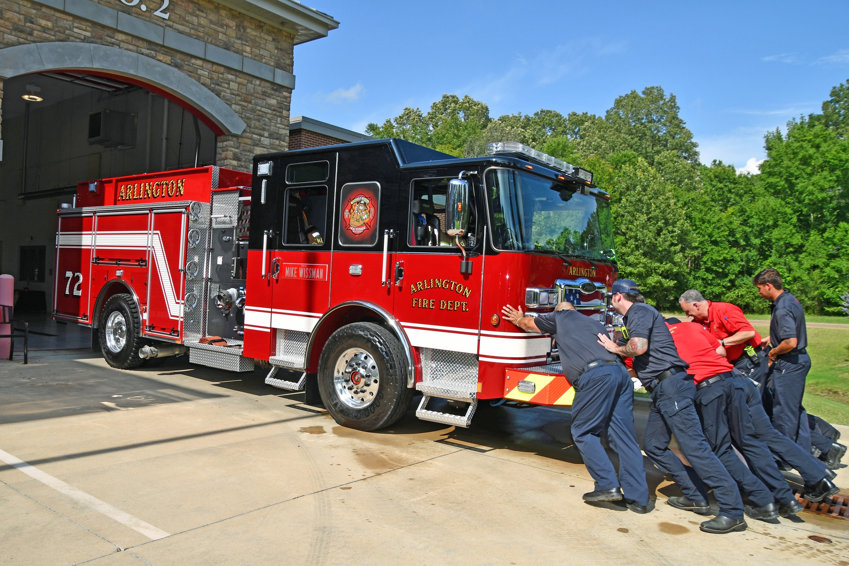 Picture of multiple fire personnel pushing a red and black fire engine, labeled as Arlington Fire Department 72, into a Fire station bay.