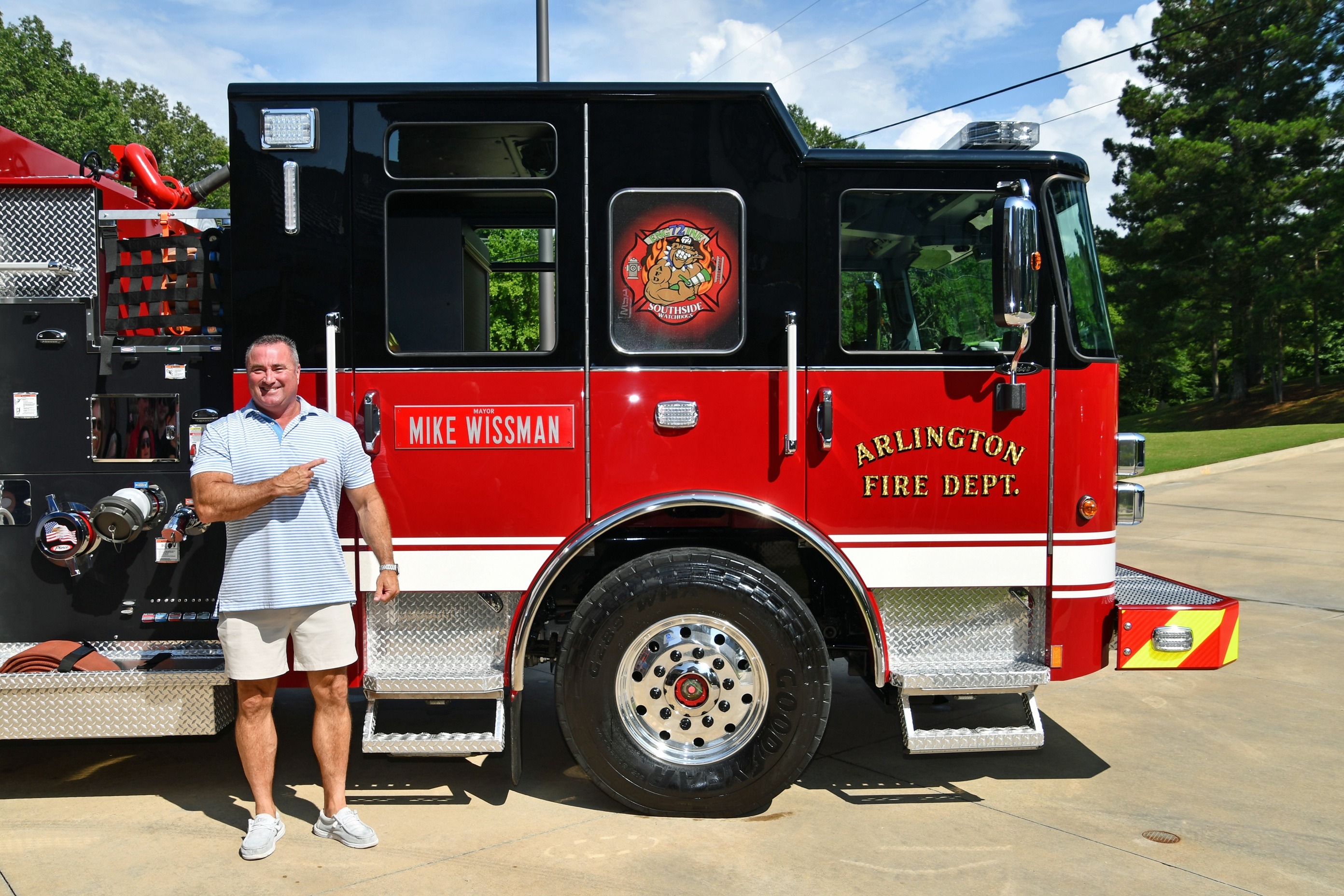 Picture of man in khaki shorts and blue polo posing in front of black and red fire engine with label of Arlington Fire Department and plaque on rear door that says Mayor Mike Wissman