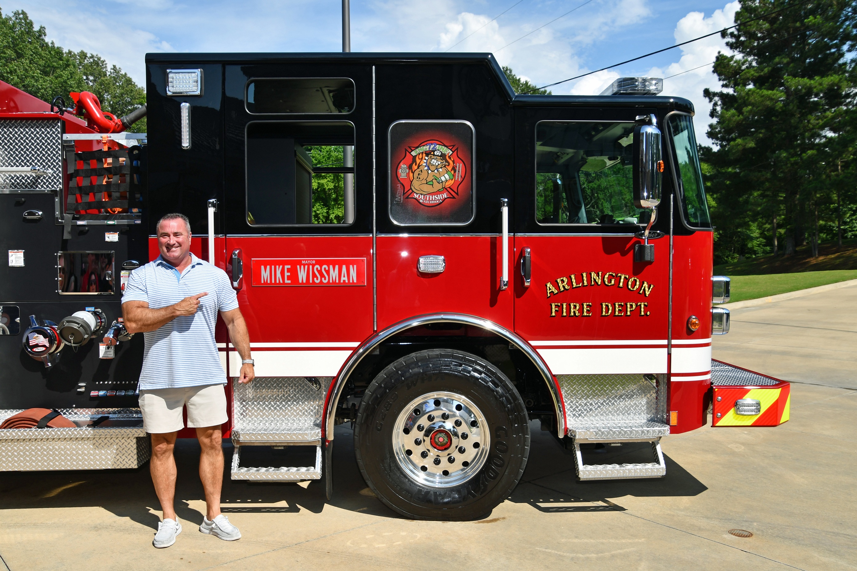 Picture of man in khaki shorts and blue polo posing in front of black and red fire engine with label of Arlington Fire Department and plaque on rear door that says Mayor Mike Wissman