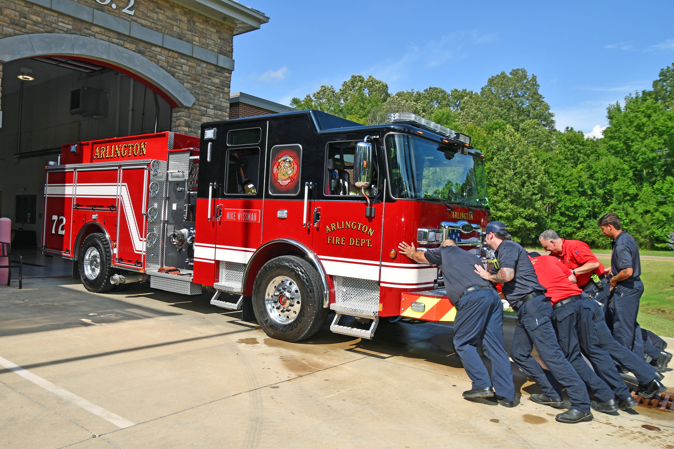 Picture of multiple fire personnel pushing a red and black fire engine, labeled as Arlington Fire Department 72, into a Fire station bay. 