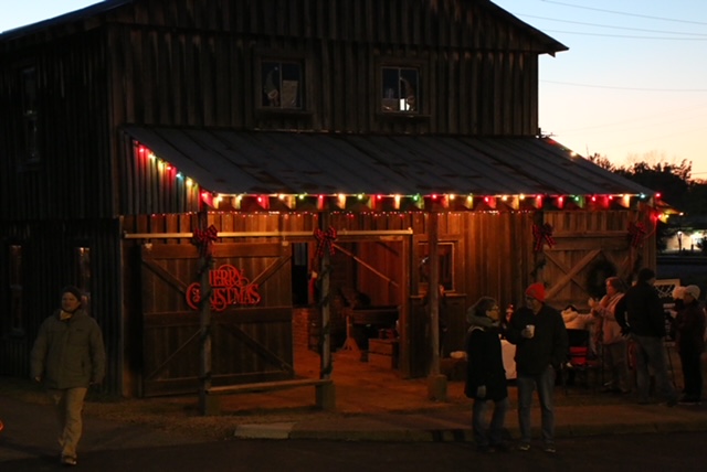 Picture of wooden barn and blacksmith shop at night, with multi-color holiday lights around the front awning and people in jackets and hats walking around