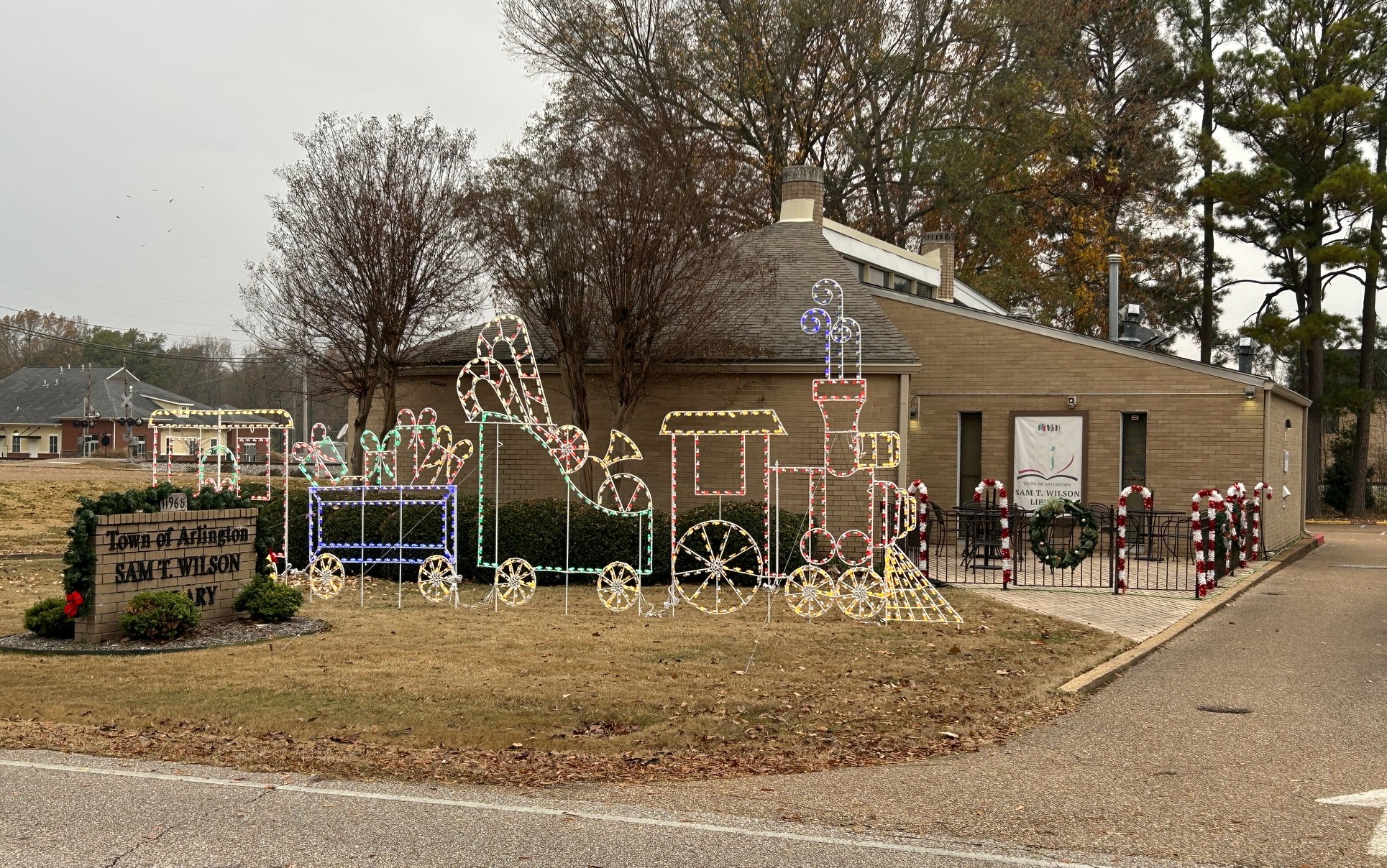 Light beige brick building with Town of Arlington Sam T Wilson Library sign out front and holiday decorations, including a holiday train made of lights and large candy canes around the patio fence