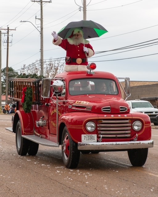 Santa Claus holding a green umbrella in the back of an antique red pickup truck and waving to a parade crows
