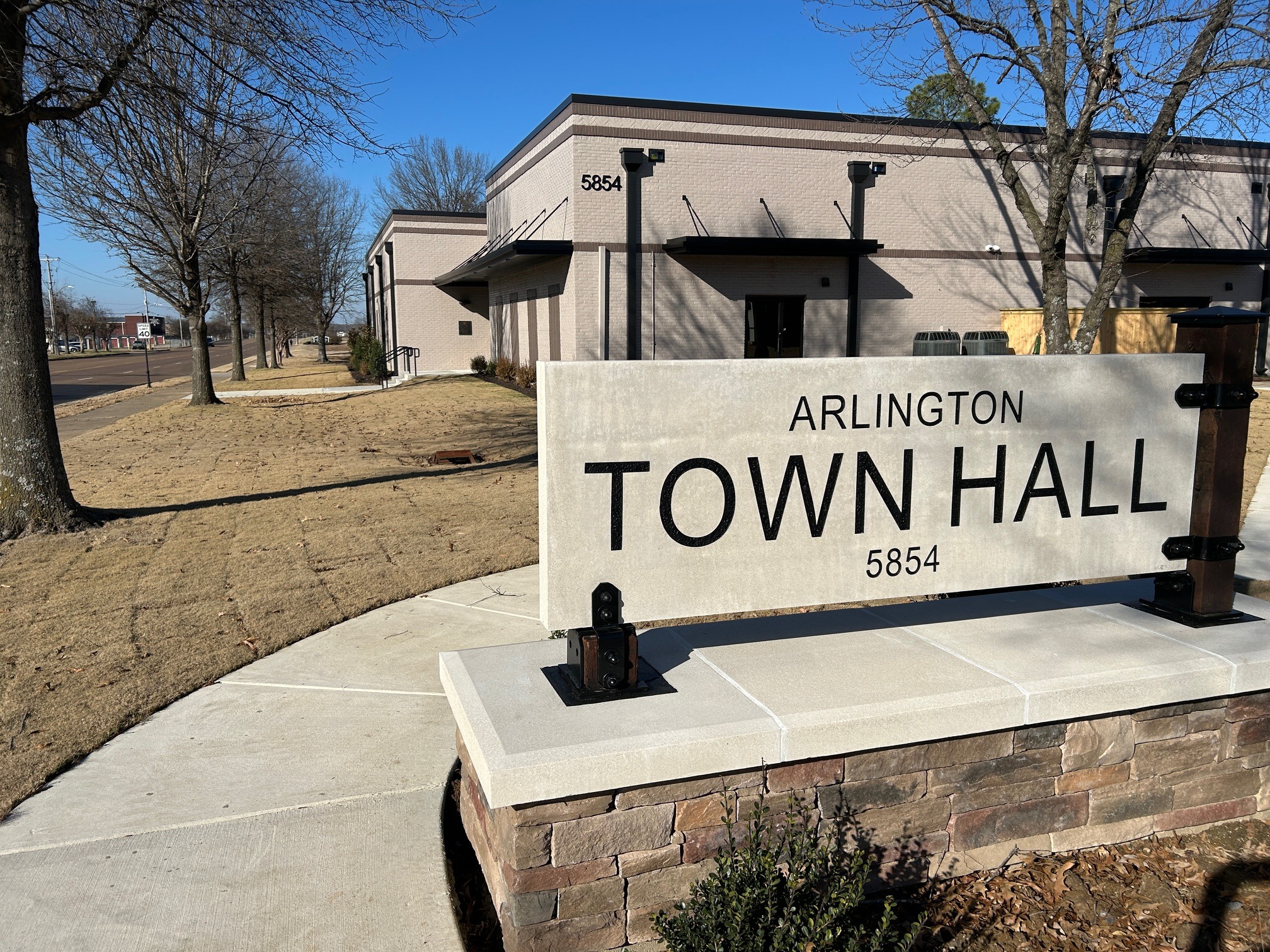 Stone monument sign with words Arlington Town Hall 5854 in brown letters. Painted brick building in background with bronze awnings and trim.