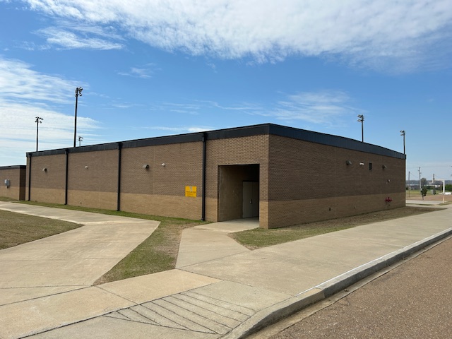 Picture of a one-story brick building with no windows. On wall by a entryway is a yellow tornado safe room sign