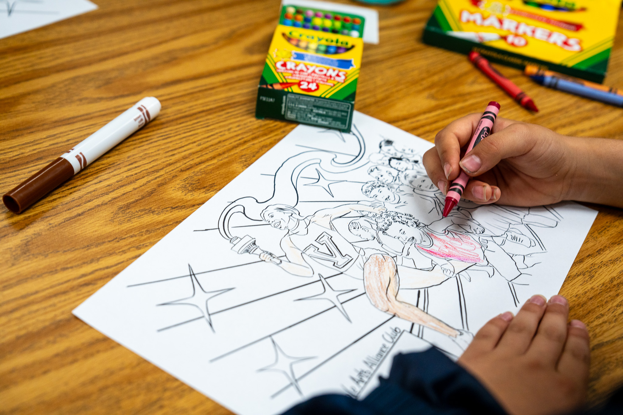 A child is coloring a large drawing on a table, with a red crayon, surrounded by a box of crayons.