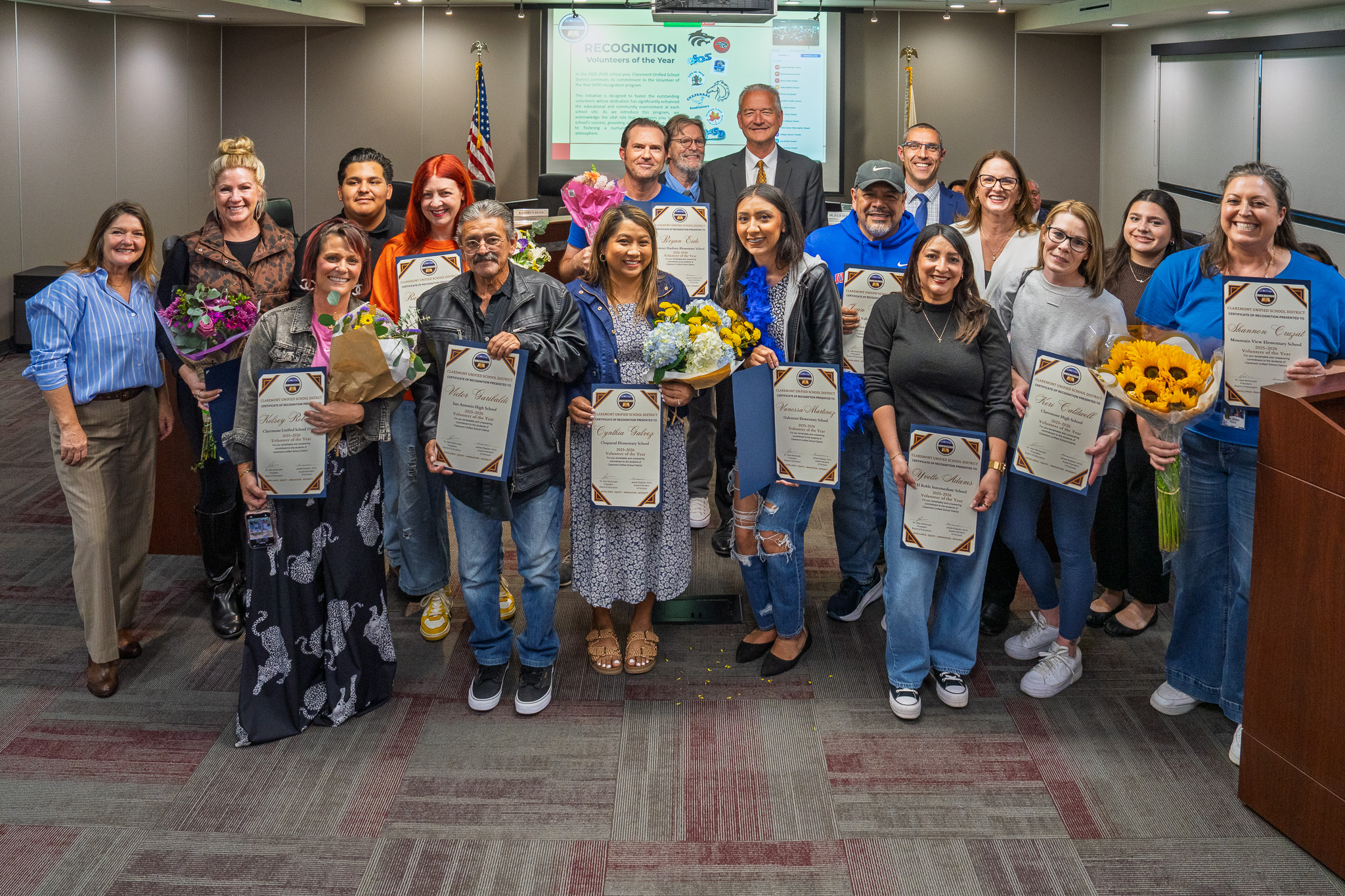 A group of adults stand in a room, some holding certificates, with a podium, a projector screen, and flowers.