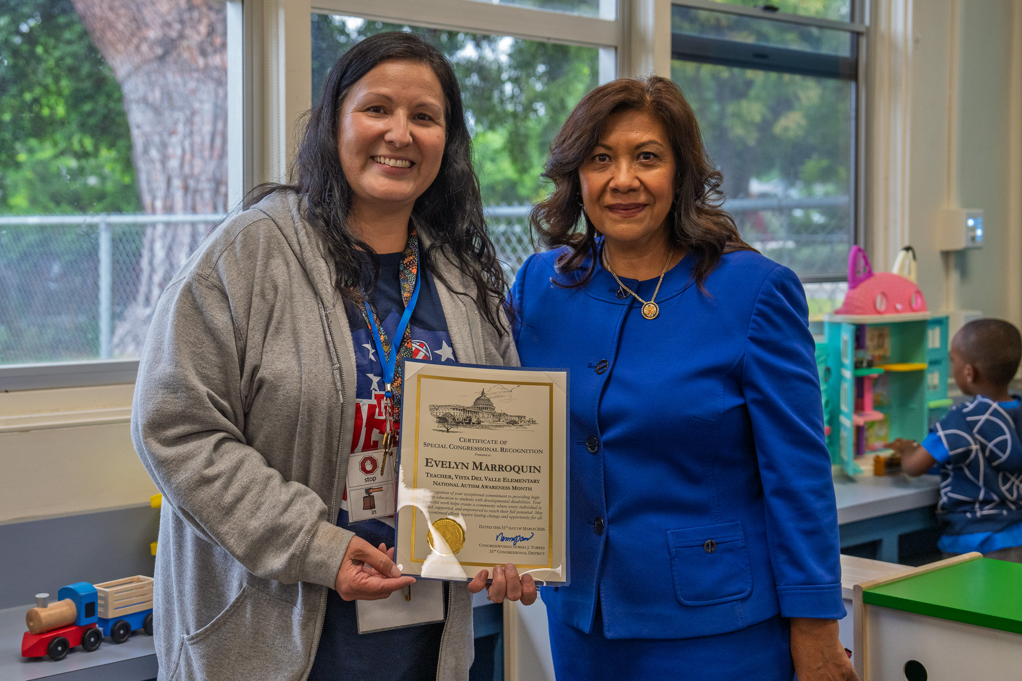 Two women hold a framed certificate; one wears a gray jacket, and the other wears a blue suit. Behind them, a child plays in a toy-filled area.