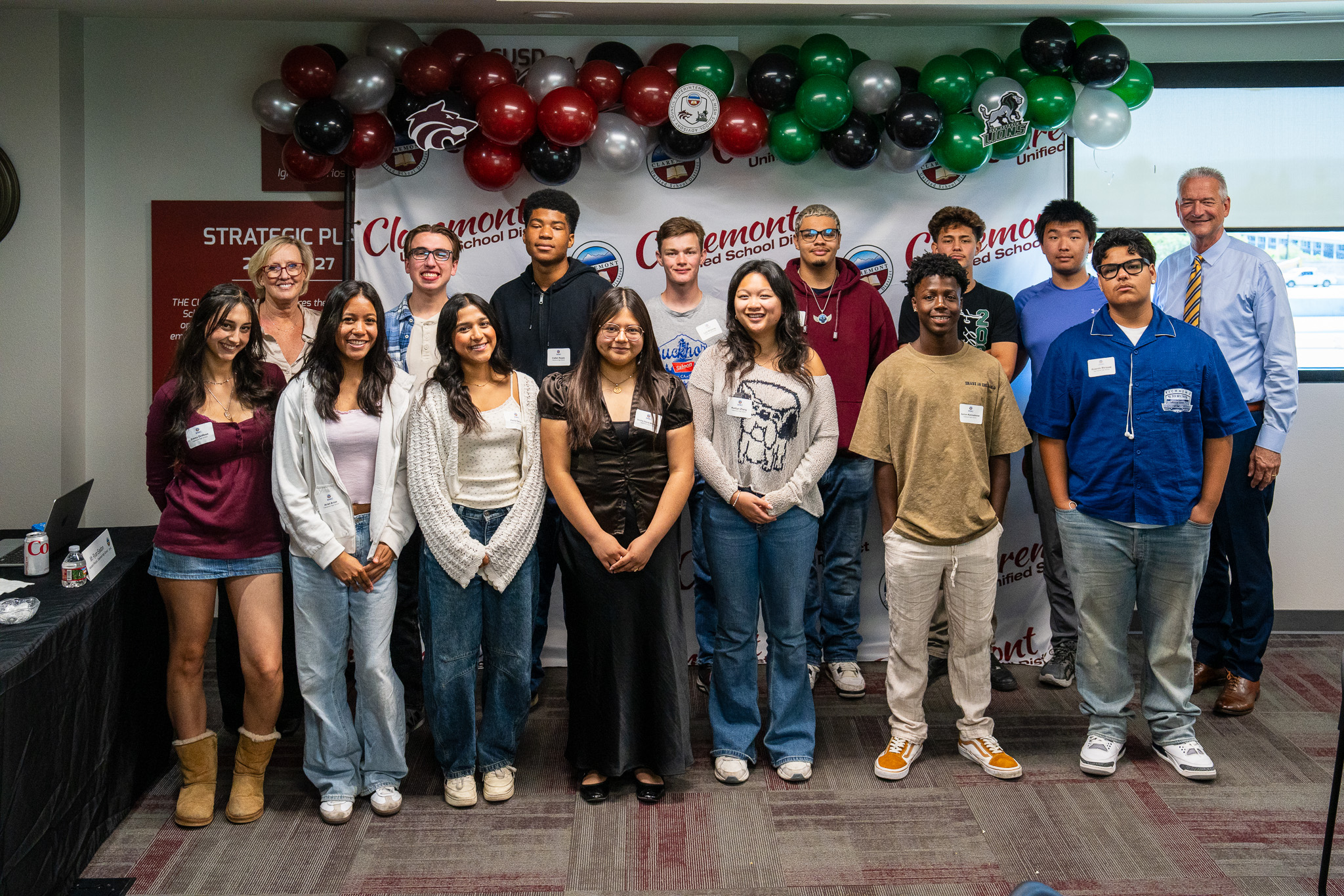 A group of young adults, some in casual clothing, stands in a line in a room. Behind them, a banner reads "Oment".