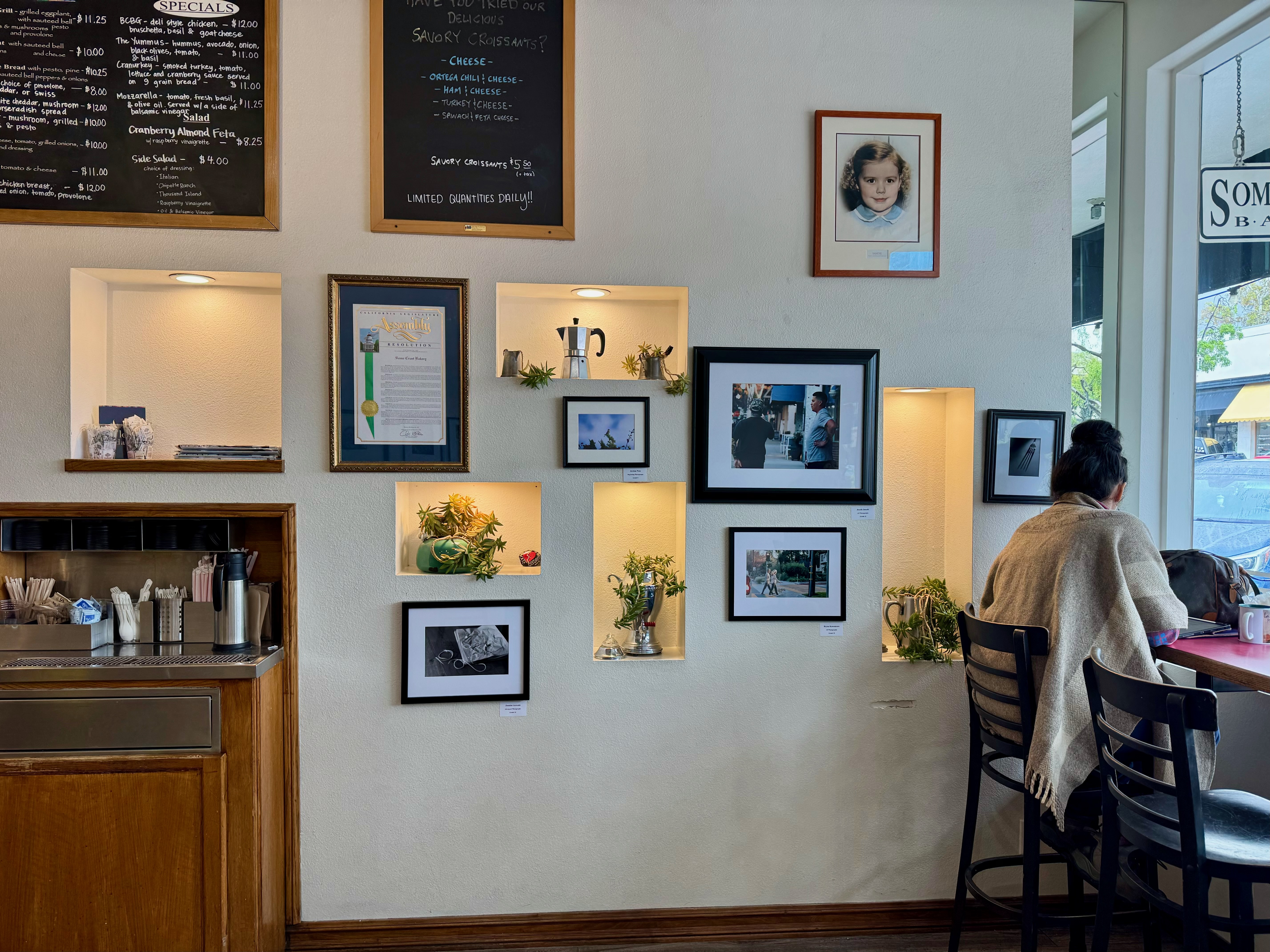 A café features a woman sitting at a counter. Frames line a wall behind her. A chalkboard sign hangs above the counter.