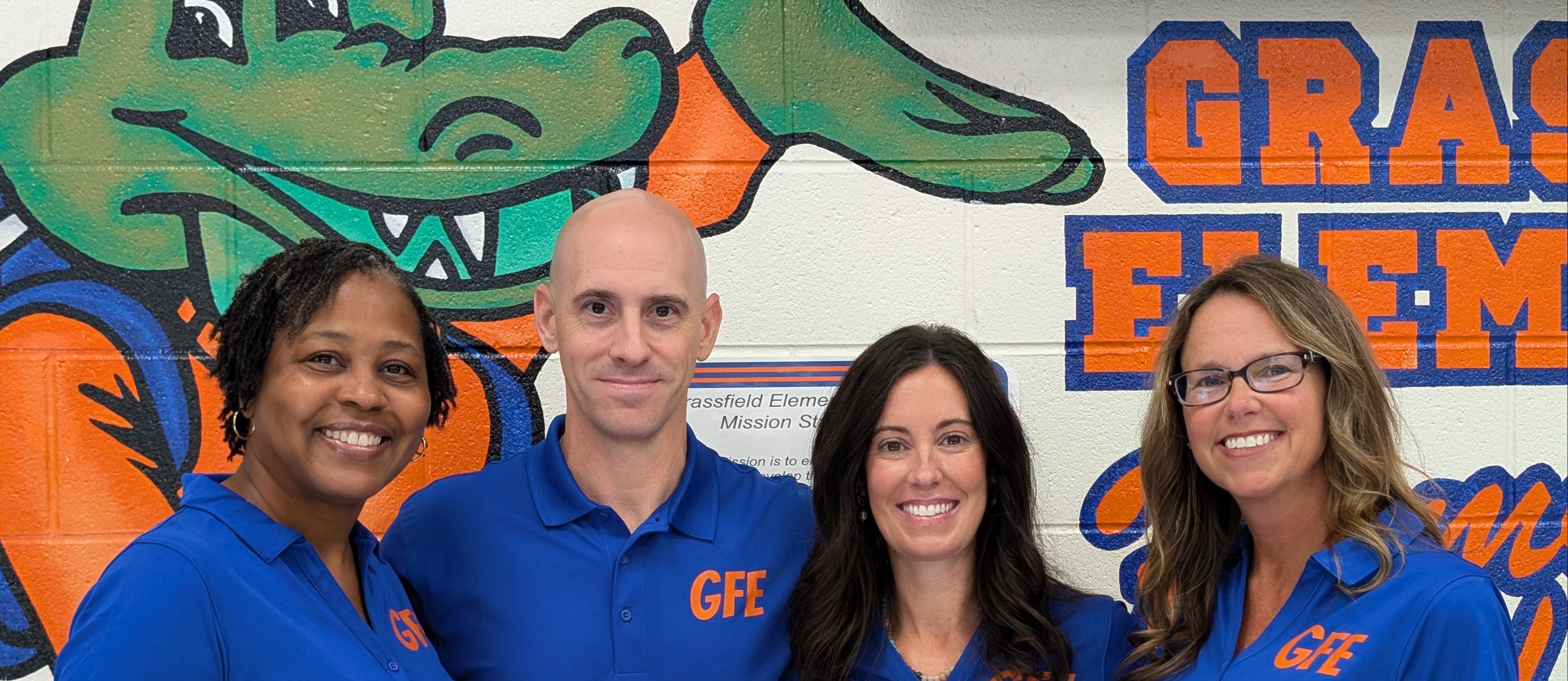 Four Grassfield Elementary staff members wearing matching blue polo shirts with "GFE" embroidered in orange, standing together in front of a school mural.