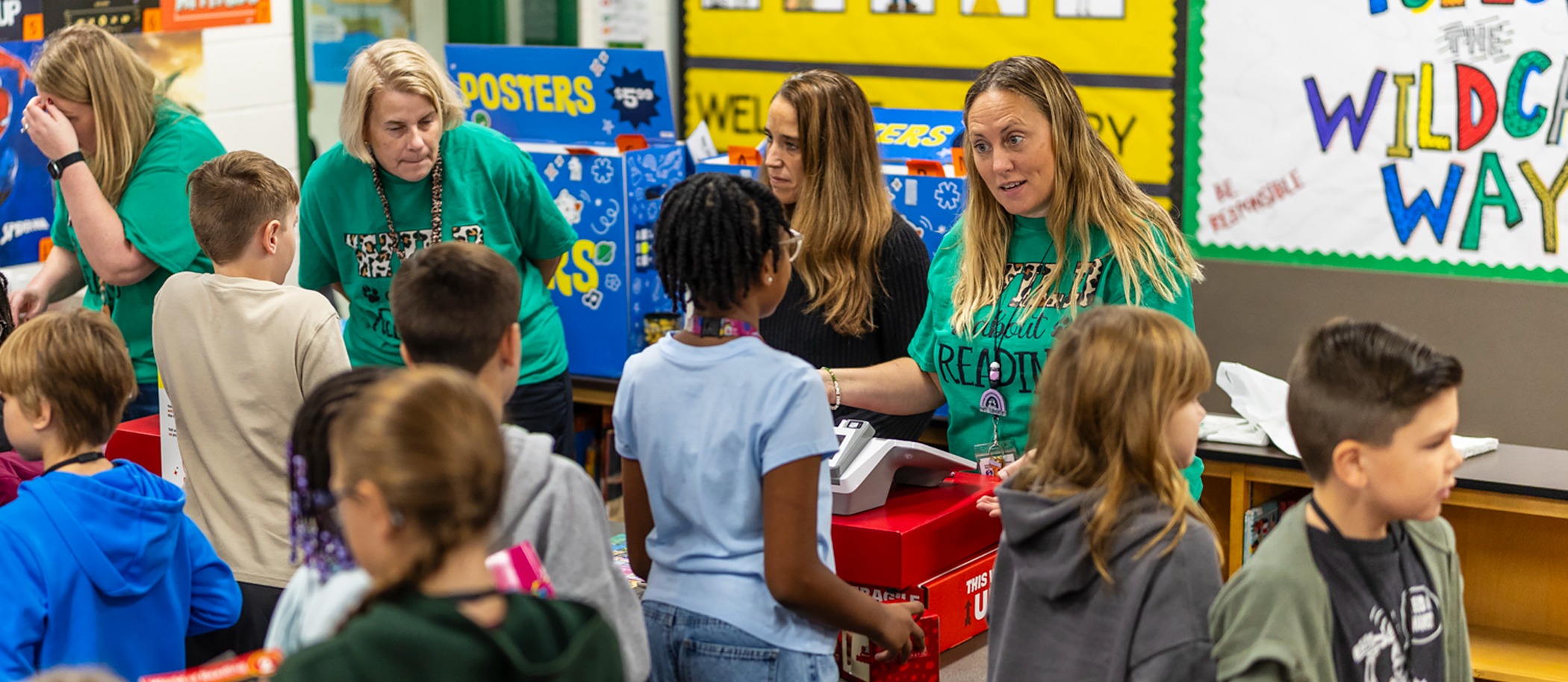 Several people, including adults and children, are standing in a classroom. Some children are standing around a table.