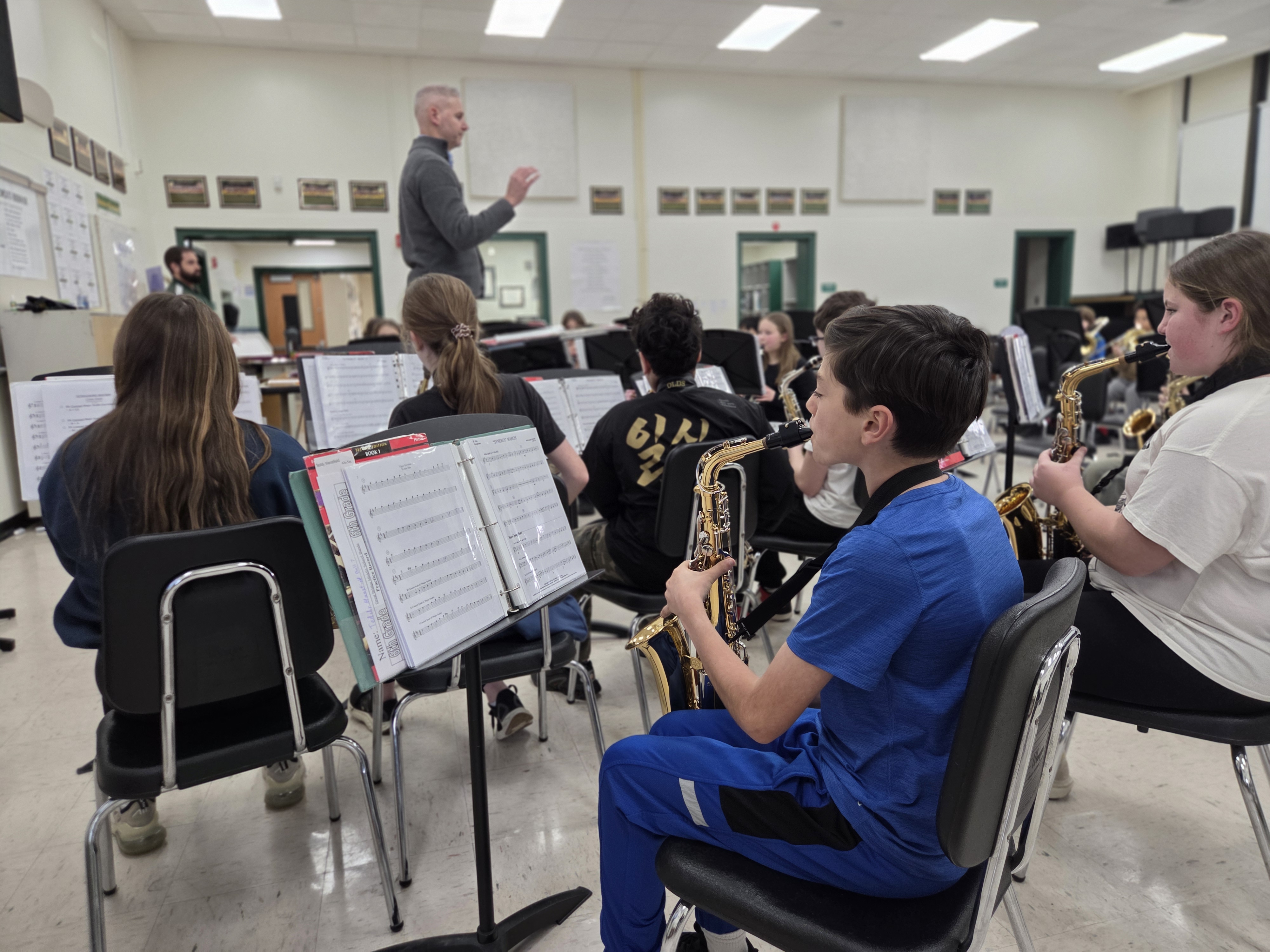 A middle school band rehearsal in a bright classroom. In the foreground, a student in blue plays an alto saxophone while focused on his sheet music. In the background, a conductor leads the ensemble of young musicians.