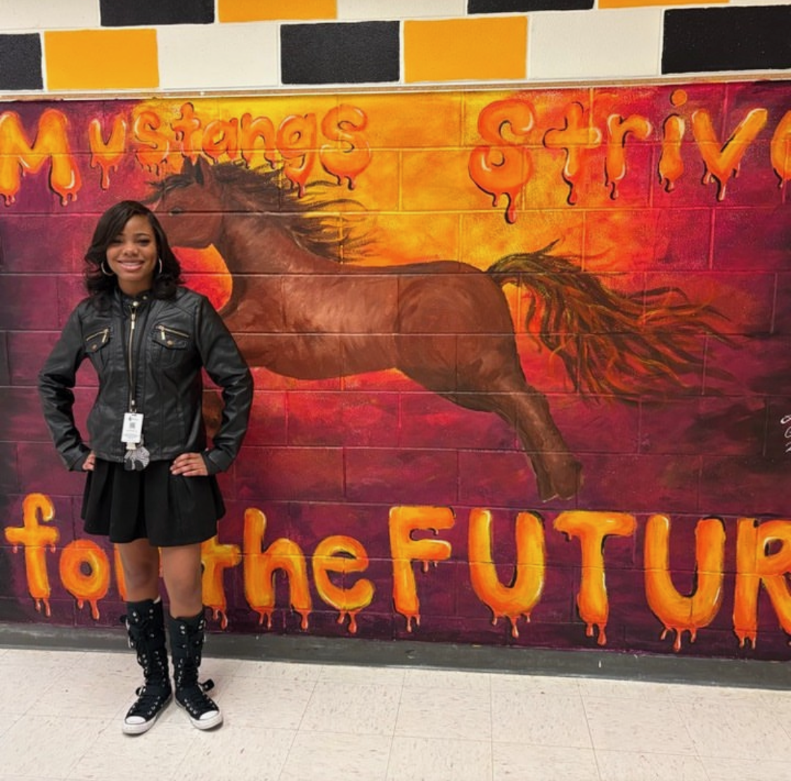 Artist standing in front of the mural she designed.  A galloping horse on a yellow to red gradient background with the words "Mustangs Strive for the Future."