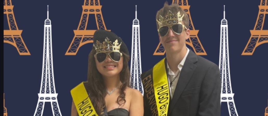 A smiling boy and girl posing together against a dark blue backdrop patterned with orange and white Eiffel Towers. The girl wears a black strapless gown, a beret with a gold crown, sunglasses, and a yellow sash reading "HUGO 8TH GRADE PRINCESS". The boy wears a black suit, a white shirt, a gold crown, sunglasses, and a yellow sash reading "HUGO 8TH GRADE PRINCE ENDEAVORS PRINCE".