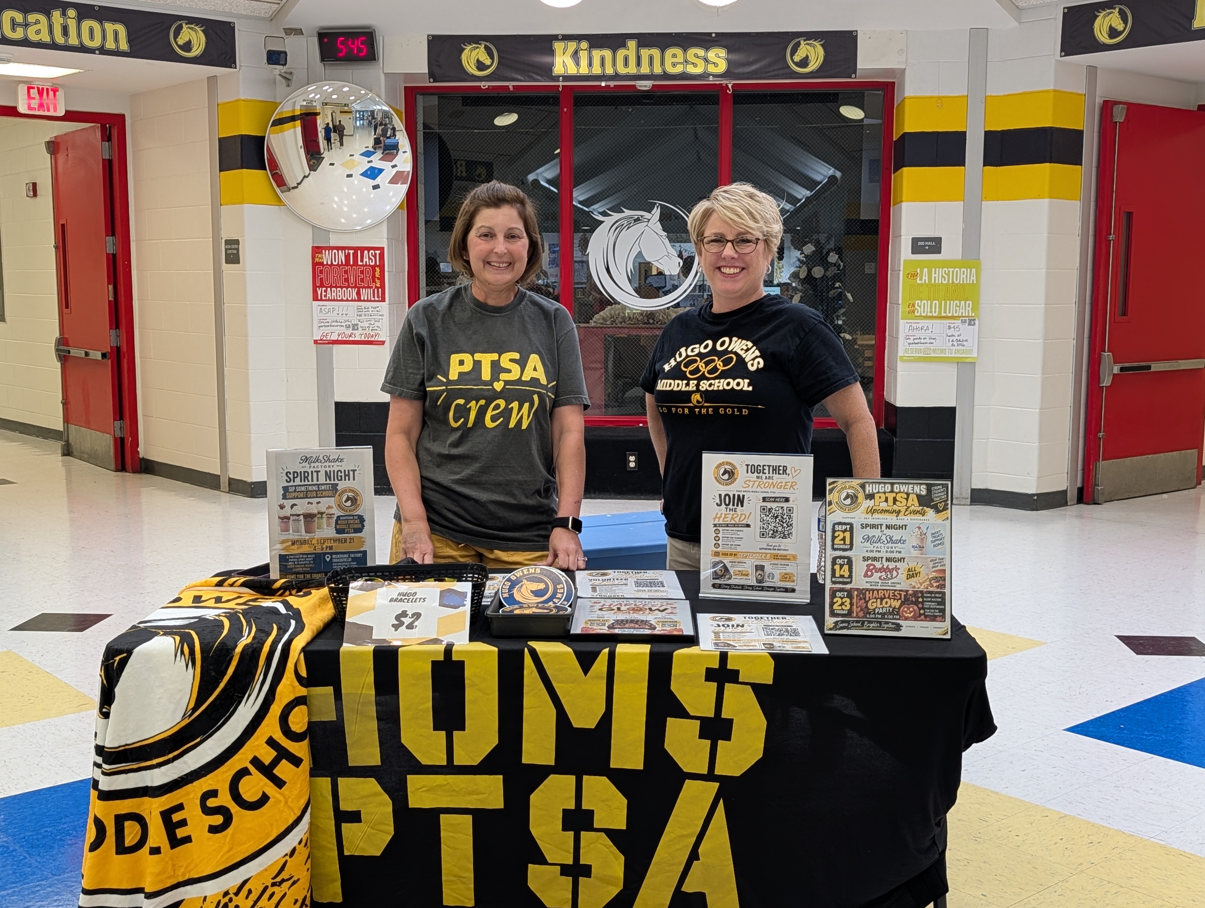 Two smiling women stand behind a school table with a black tablecloth reading "HOMS PTSA" in yellow text. One wears a gray shirt reading "PTSA Crew," and the other wears a black shirt reading "Hugo Owens Middle School." The table displays promotional signs for PTSA events, a flyer reading "Join the Herd!", and a sign advertising Hugo Bracelets for $2. Above them, a sign on the hallway wall reads "Kindness."