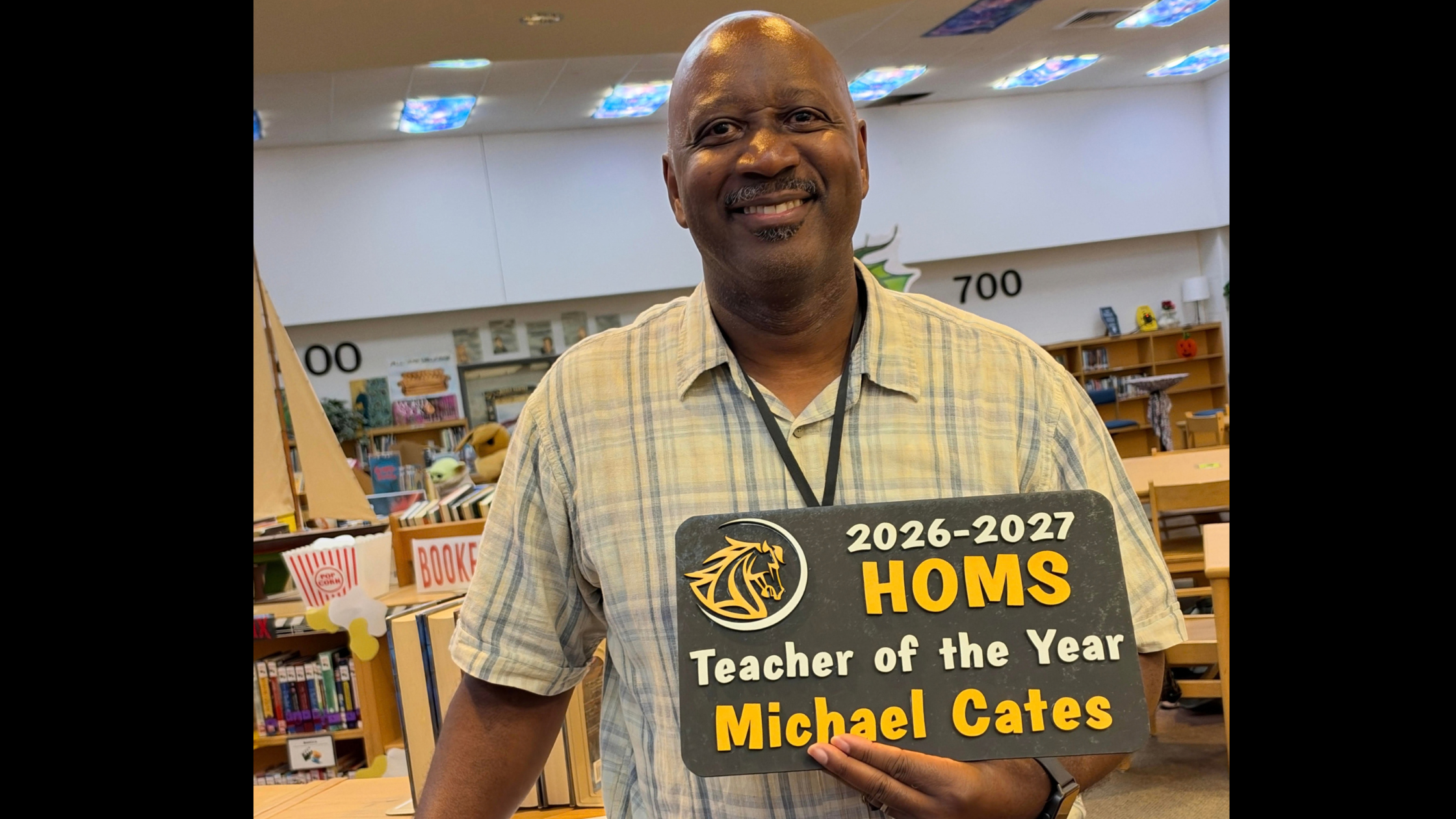 A smiling man standing in a library while holding a sign that reads, "2026-2027 HOMS Teacher of the Year Michael Cates" next to a mustang logo. 