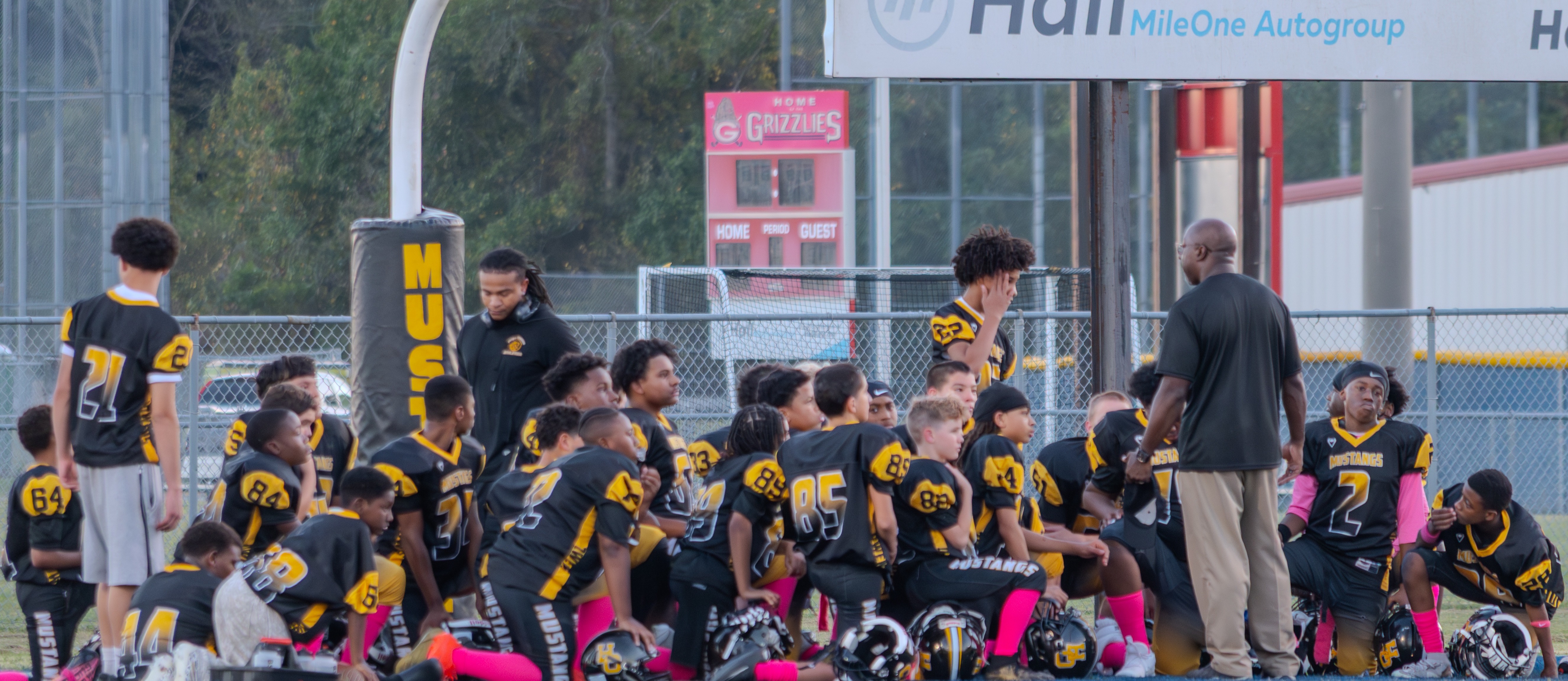 The Hugo Owens Middle School football team wearing black and yellow "Mustangs" jerseys with pink socks kneels on the sideline during a huddle during the Pink Out Game. A coach standing in black shirt and tan pants addresses the players. Positioned under the goalposts, the background shows a stadium scoreboard with sponsored signage reading "Hall MileOne Autogroup" and a pink "Home of the Grizzlies" sign in the distance.   