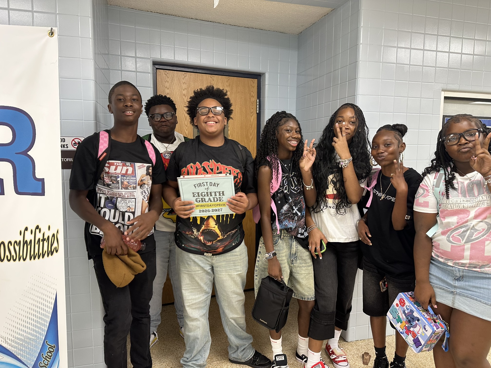 A group of seven middle school students posing together in a hallway, with a student in the center holding a "First Day of Eighth Grade 2026-2027" sign.