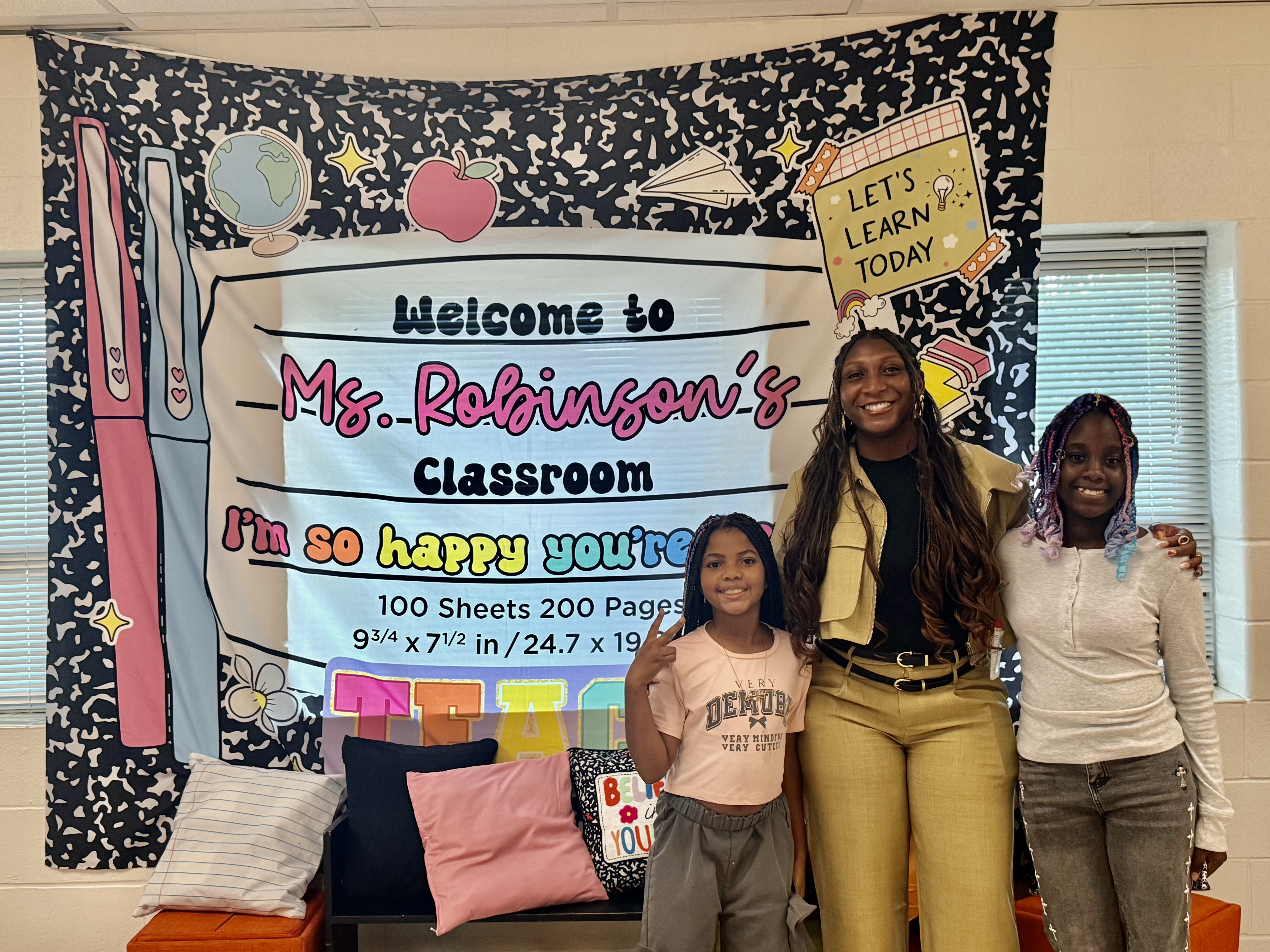 A teacher and two young students smiling and posing together in front of a backdrop designed like a composition notebook that reads "Welcome to Ms. Robinson's Classroom."