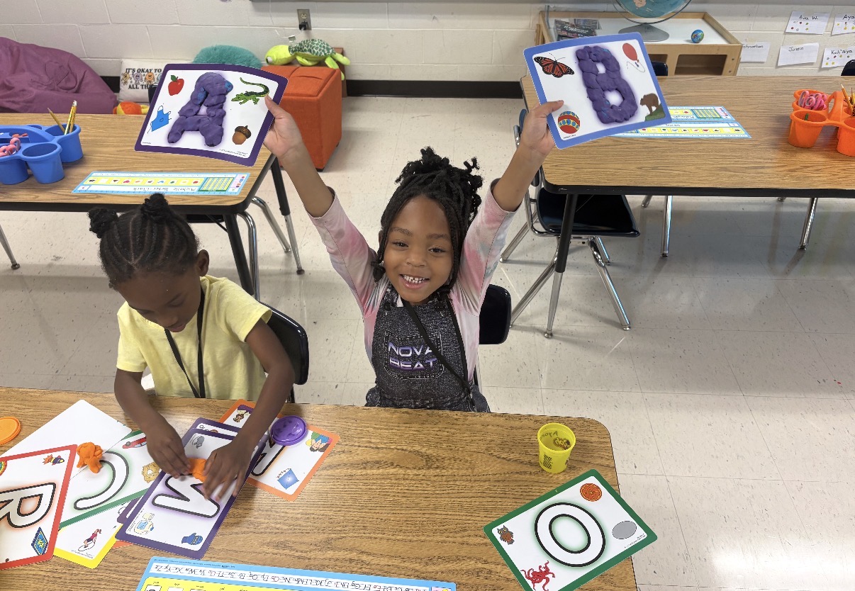 A young girl sits smiling enthusiastically at a wooden classroom table, holding up two colorful activity mats with purple playdough shaped into the letters "A" and "B". Next to her, another young girl works on a playdough letter activity on the same table.