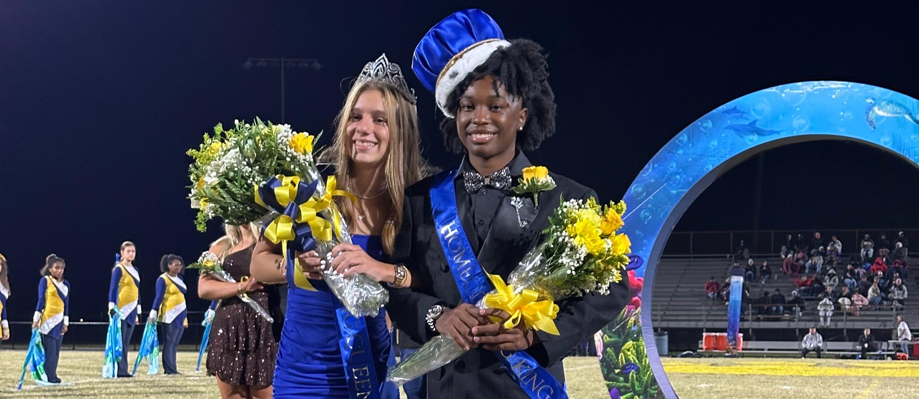 Homecoming king and queen standing on a football field, holding flowers and wearing crowns and sashes.