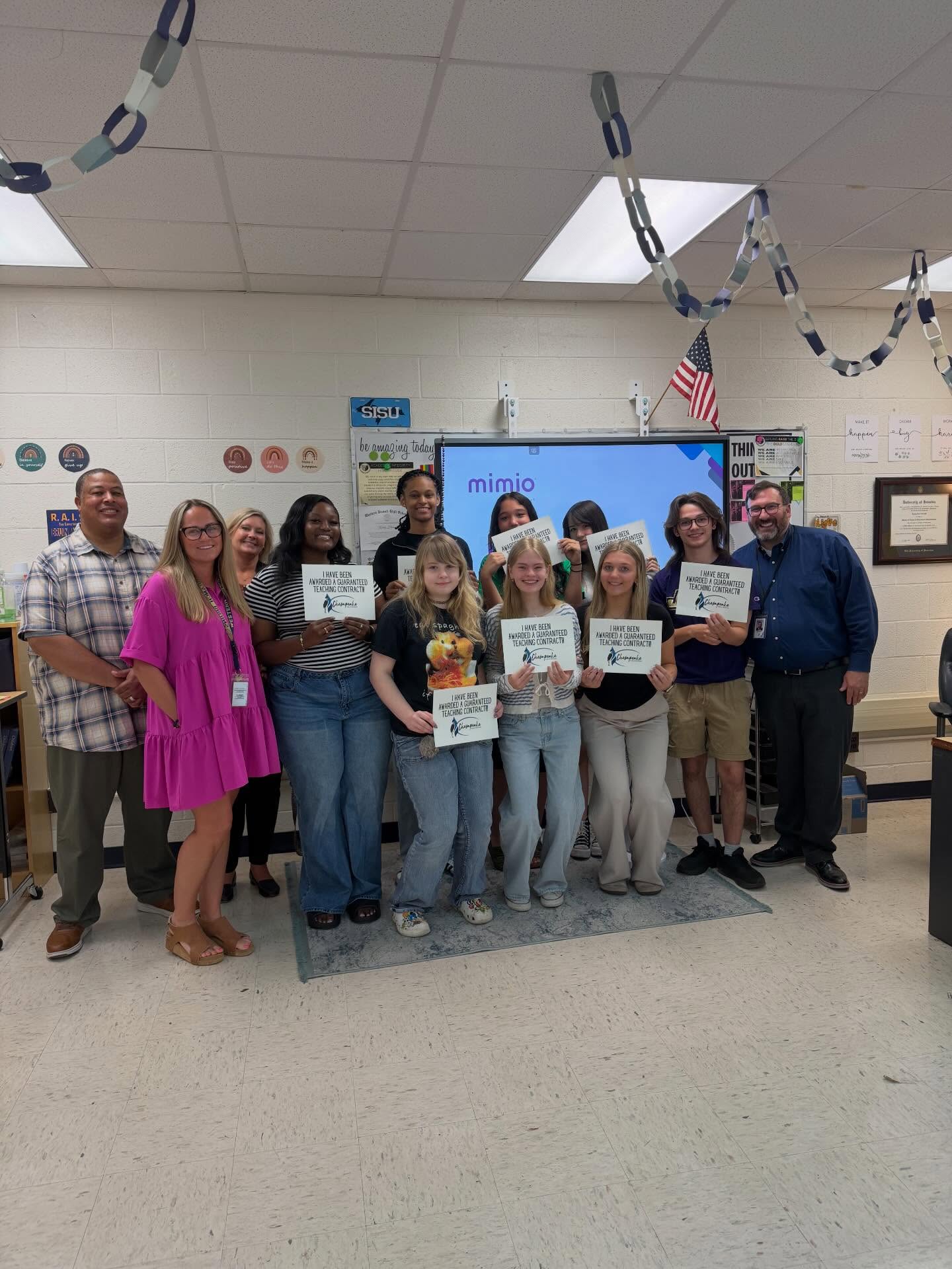 A group of students and their teacher pose together in a classroom, smiling in front of a smart board and classroom decorations.