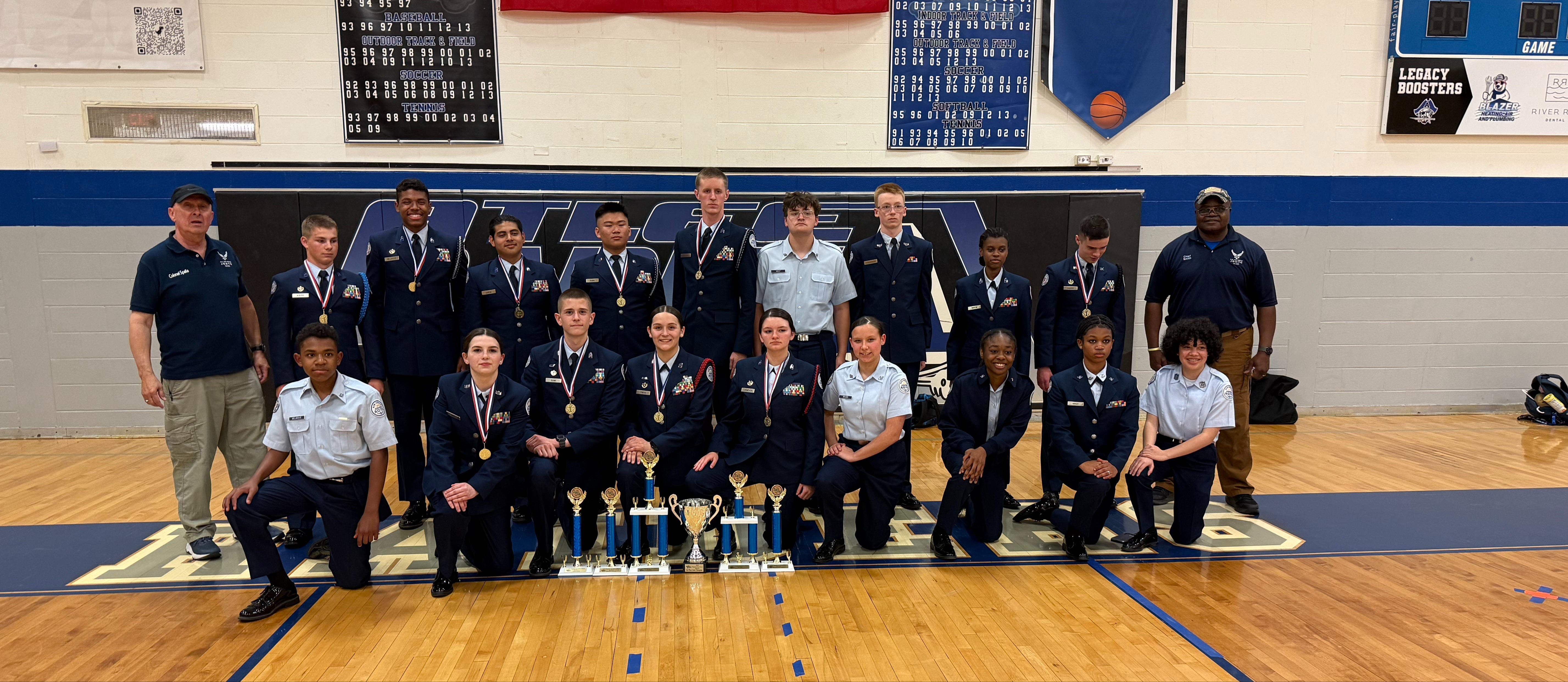 Group photo of JROTC students in military-style uniforms standing and kneeling on a high school basketball court. 6 trophies from the April 2026 State Drill Competition are displayed on the floor in front of the group. Two JROTC instructors are stand at each end. 