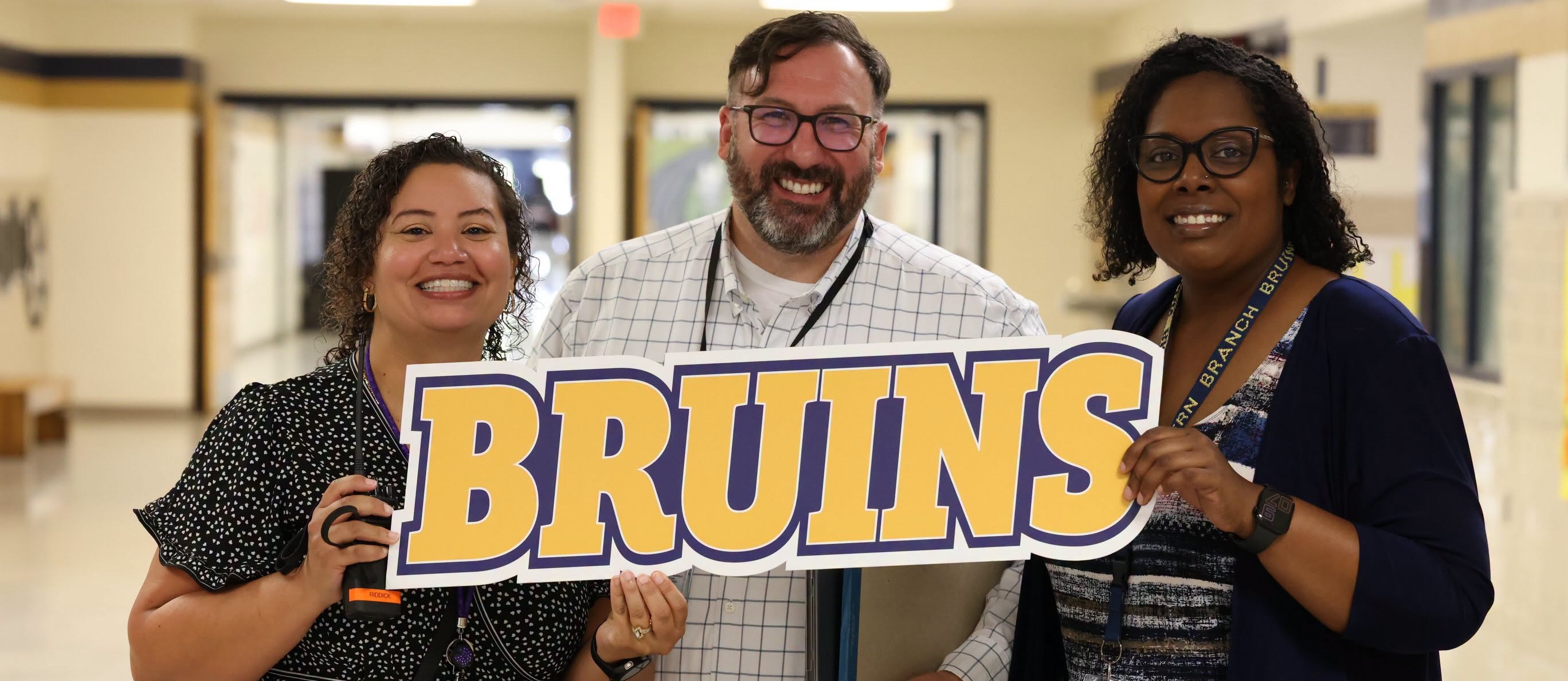 Three school staff members smile while holding a large “BRUINS” sign in a school hallway. The sign features gold letters outlined in purple and white.