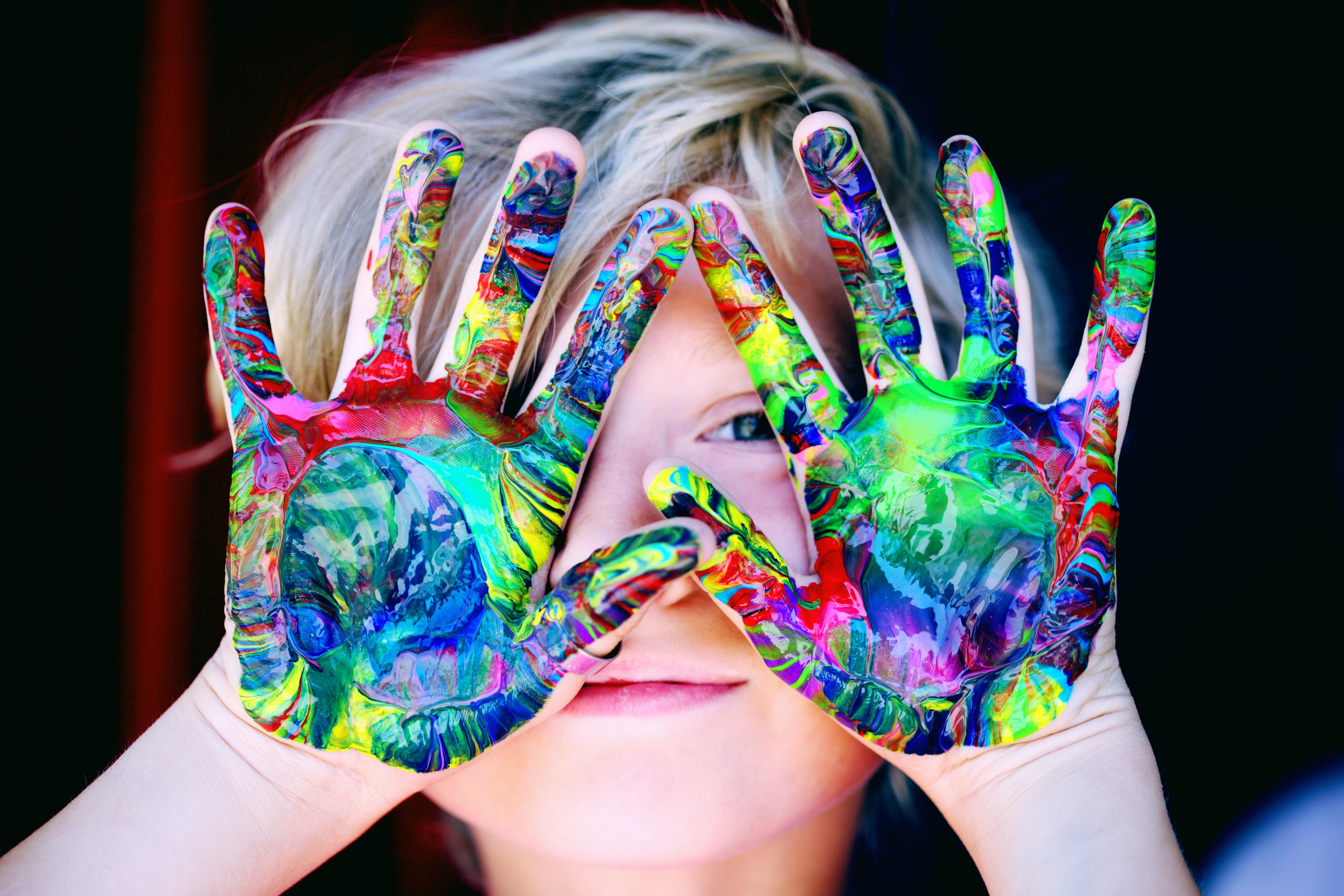 A child holding hands, painted in bright colors, positioned in front of the face, partially obscuring their face.