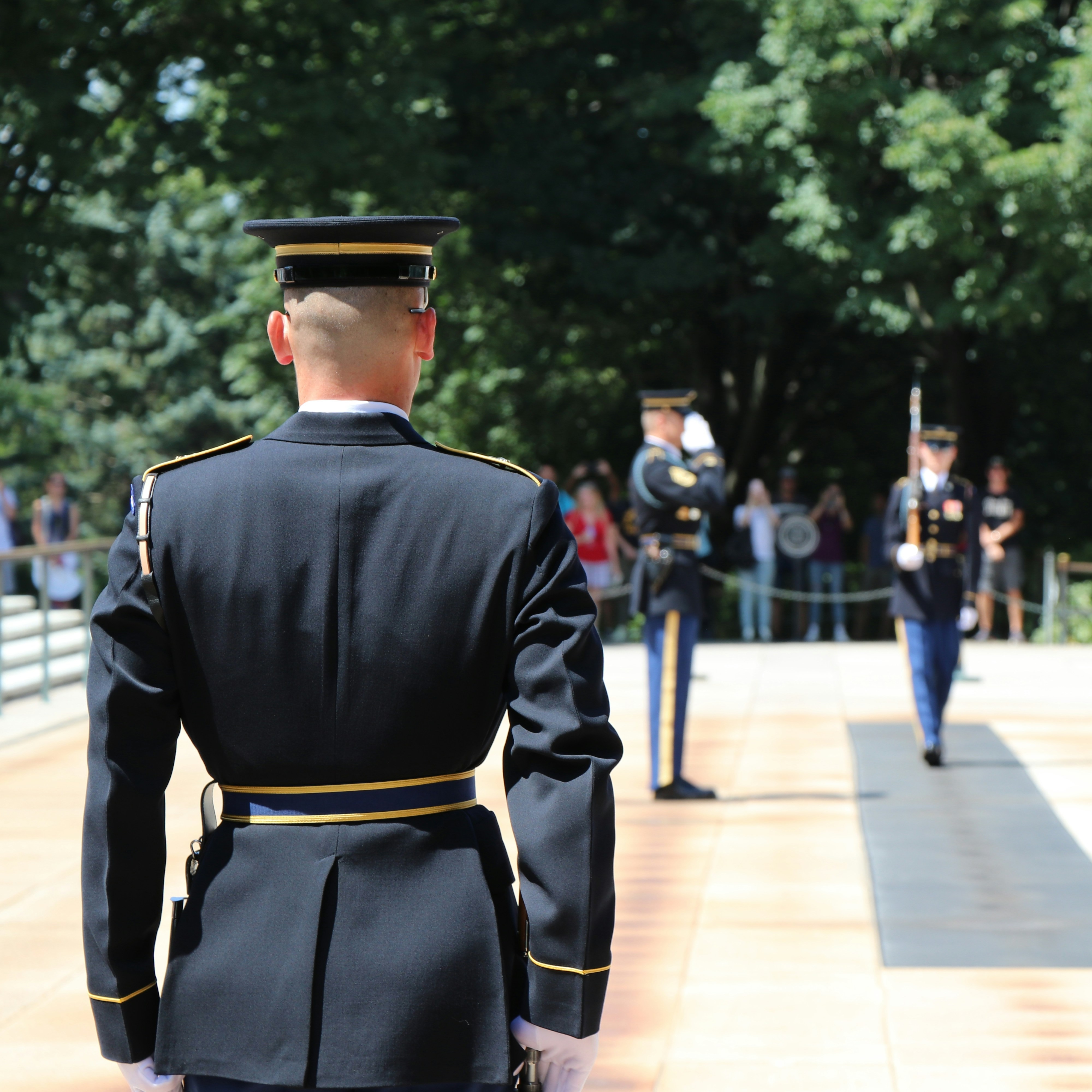 Military personnel in uniform stand outside with green trees in the background. One faces away.