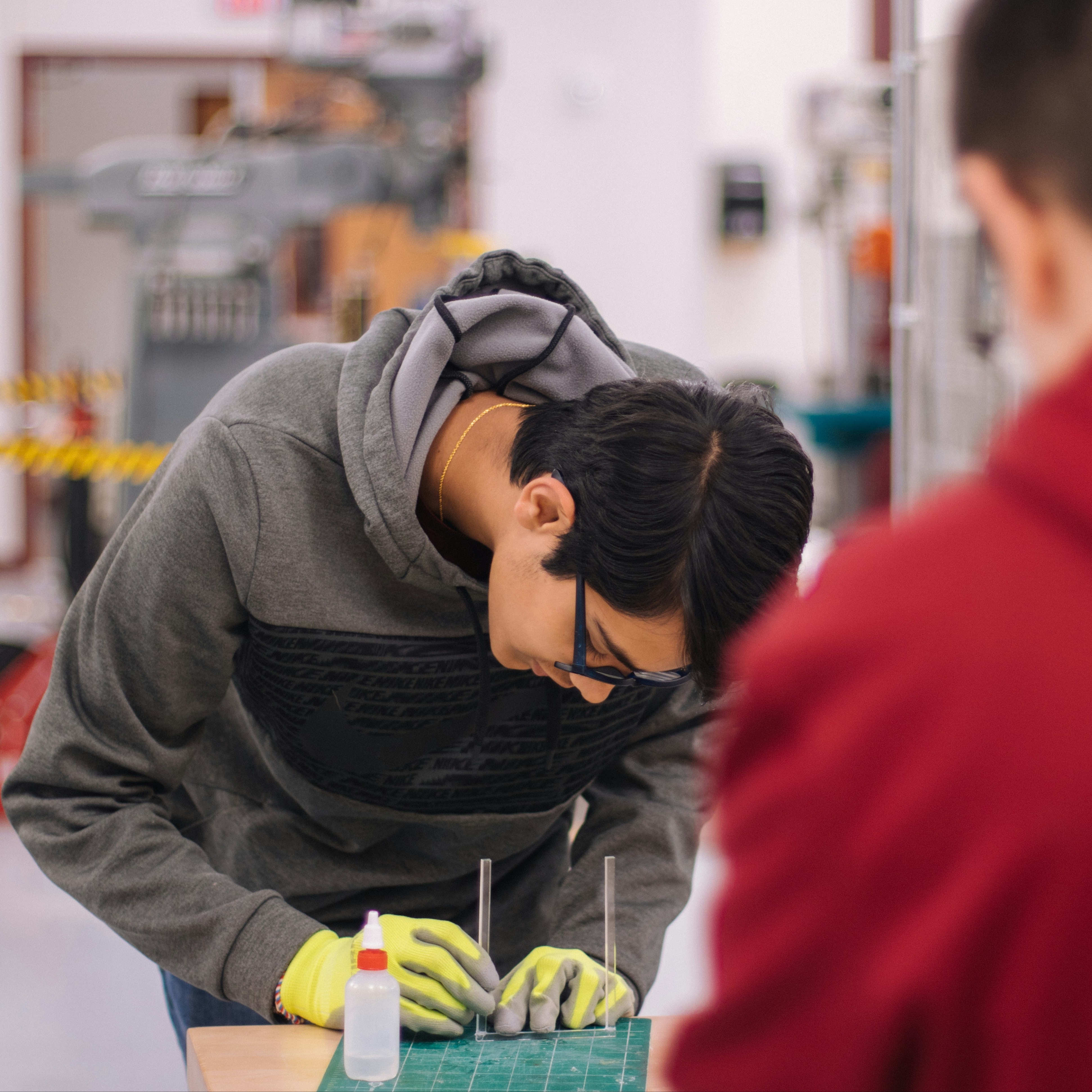 Two individuals in a workshop; one wearing a gray hoodie, yellow gloves, and glasses is working on a project.
