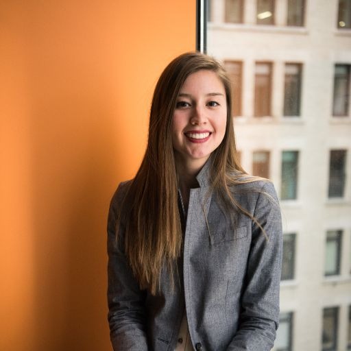 Woman with long hair wearing a gray blazer smiles in front of a building with glass windows.
