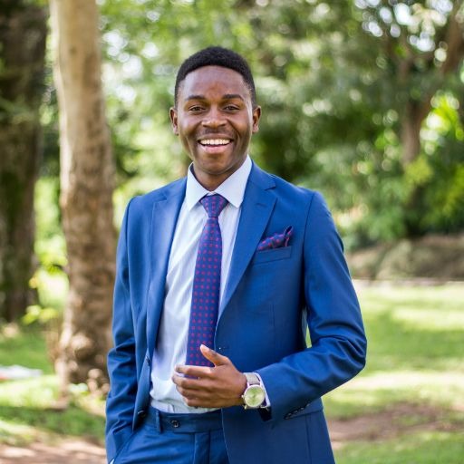 A man in a blue suit and tie smiles, hands in pockets, standing in a park with trees.
