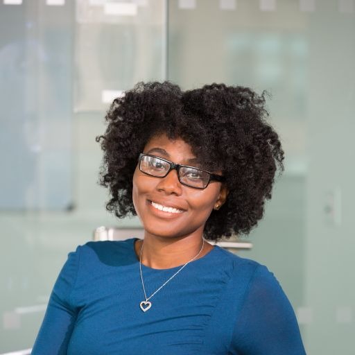 A smiling woman with glasses and curly hair in a blue blouse poses against a glass wall.