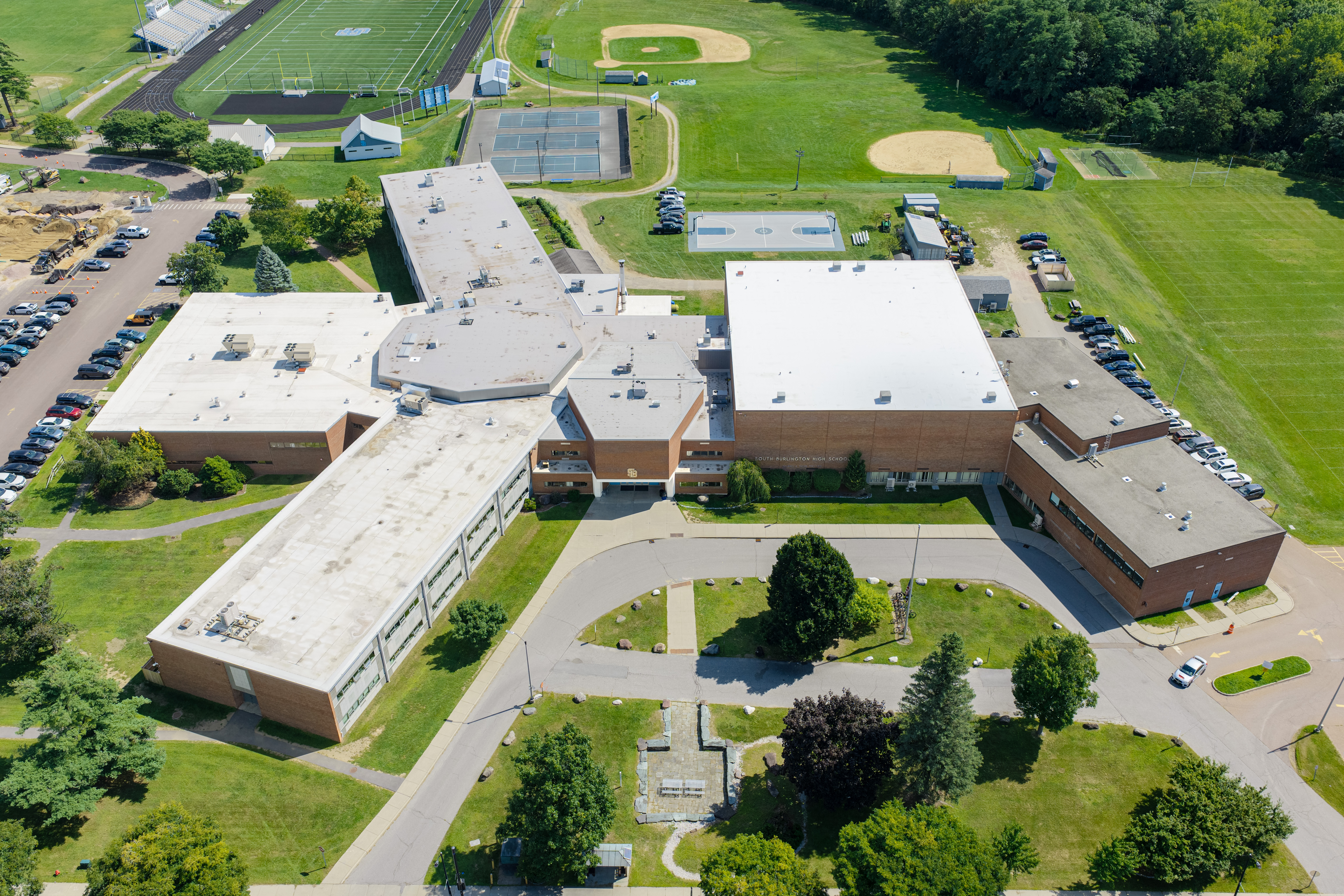 Aerial view of a school building with multiple wings, surrounded by greenery, parking lots, and a baseball field.