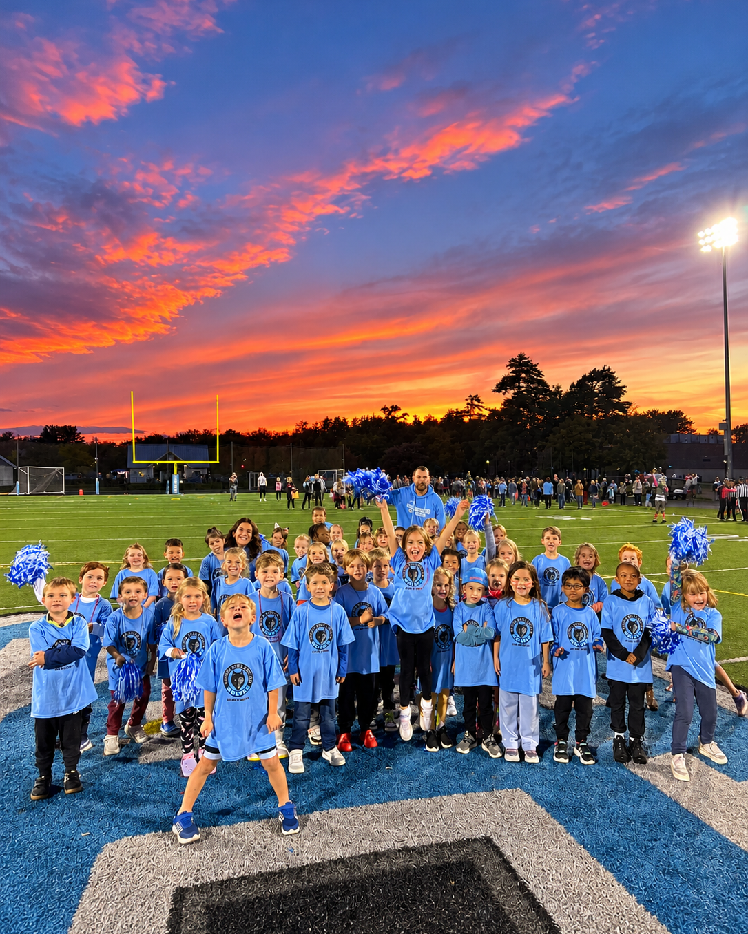 A group of young students wearing blue shirts on a football field. The sky has a sunset hue.