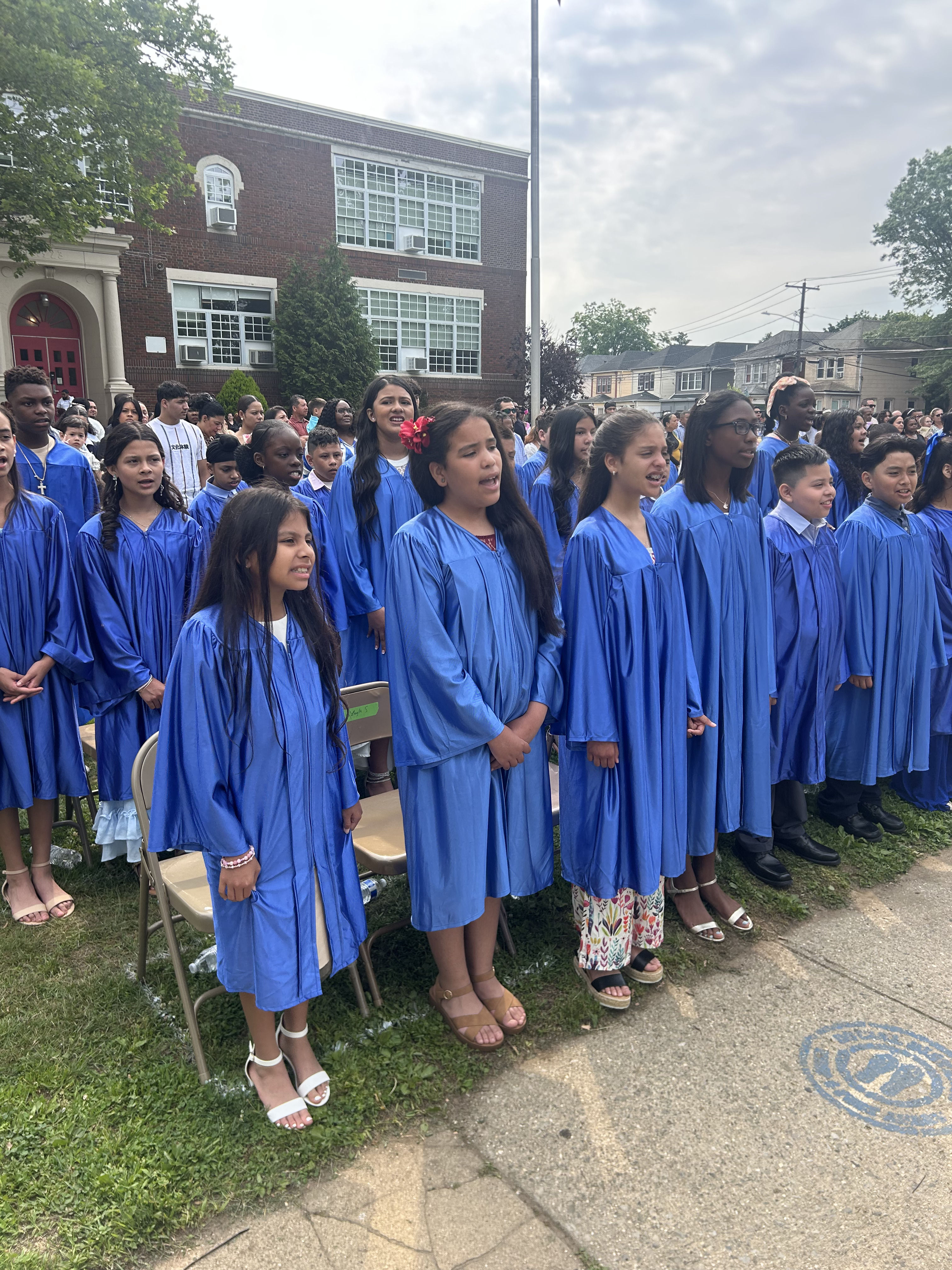 Students graduating wearing blue graduation gowns standing outside of the front of the school
