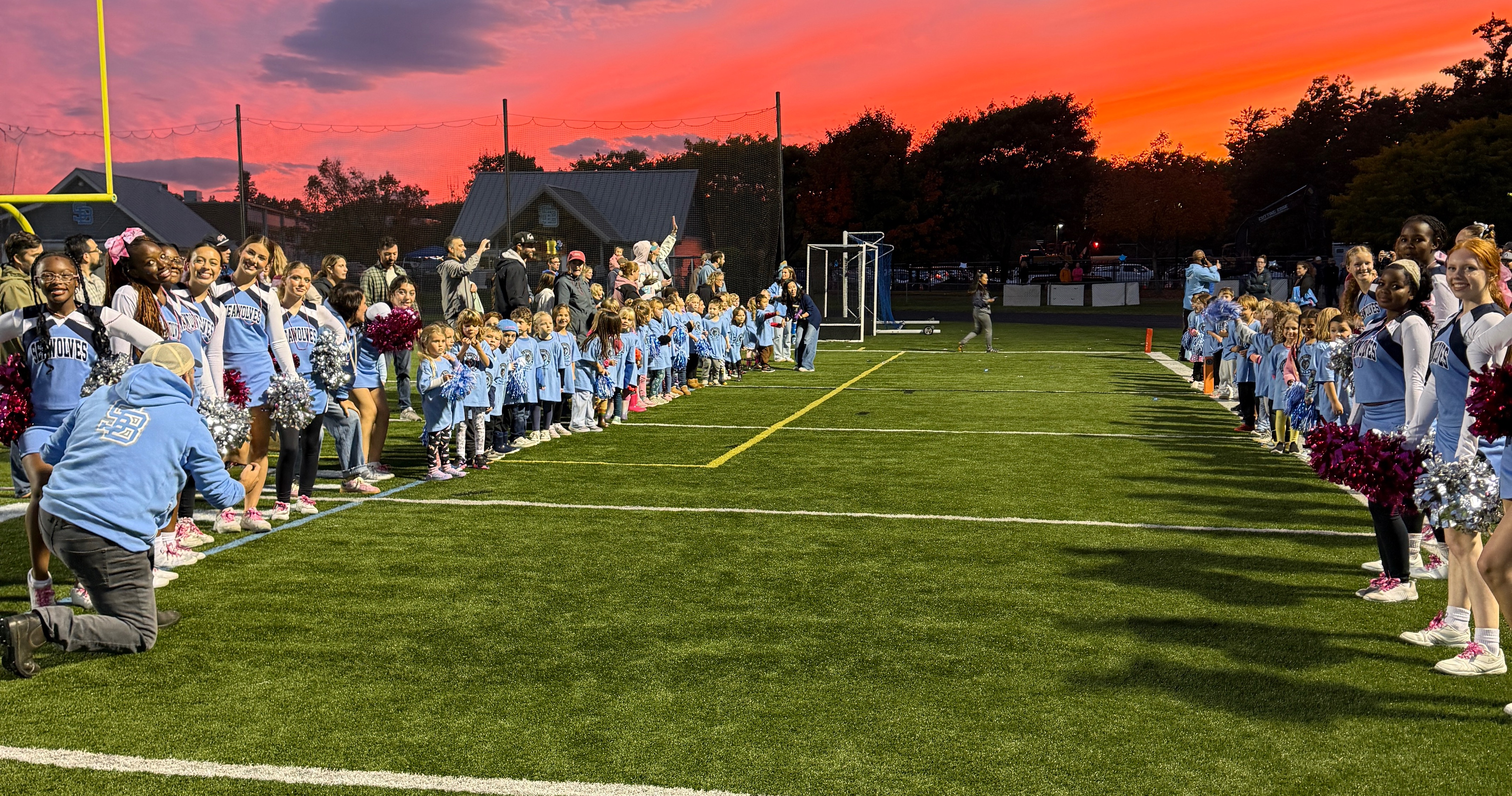 Many people dressed in blue stand in two rows on a field. One person kneels at the front.