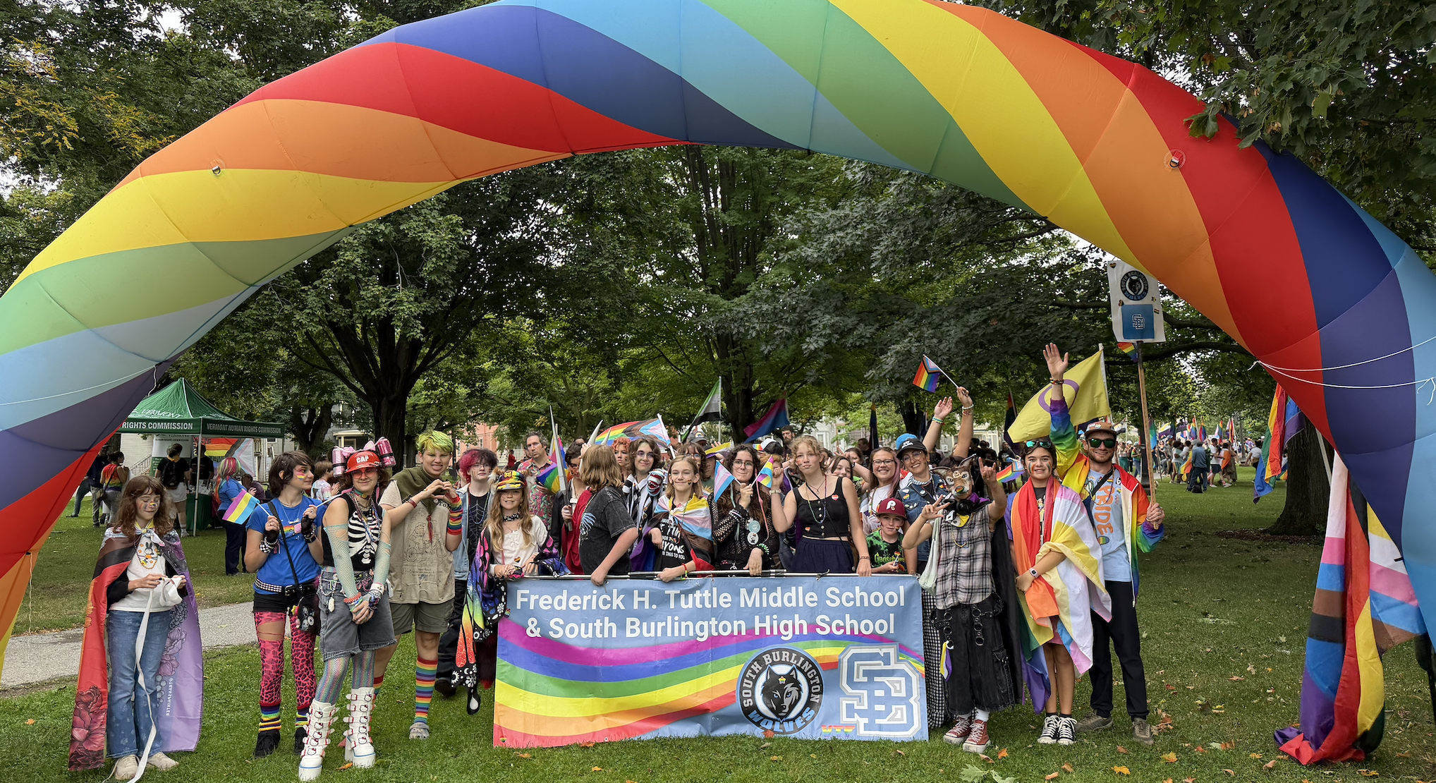 A group of people stand beneath a large, rainbow-colored arch, holding banners, and smiling.