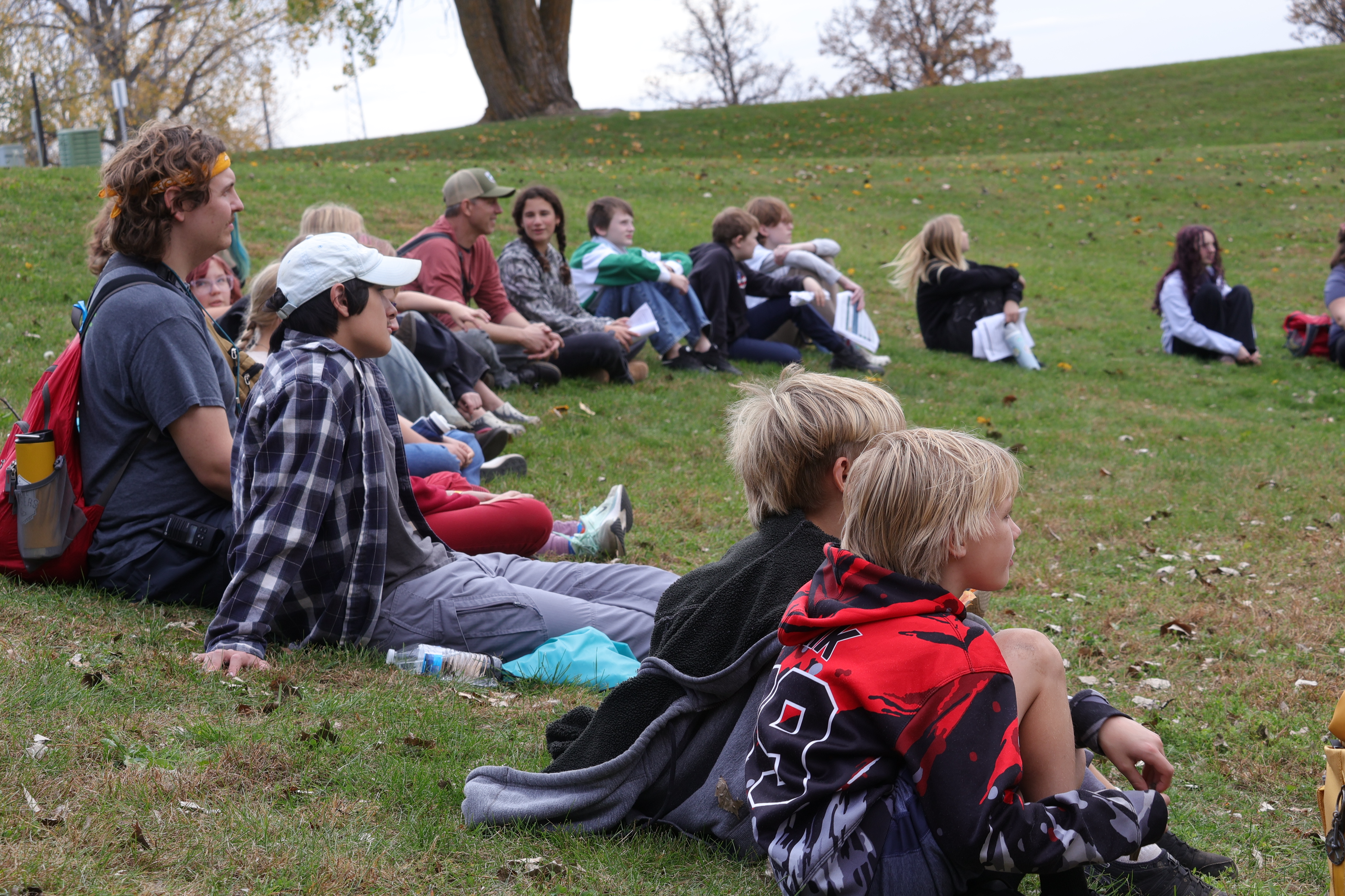 A group of people sitting on a grassy hill, some wearing hats, sneakers, and backpacks.