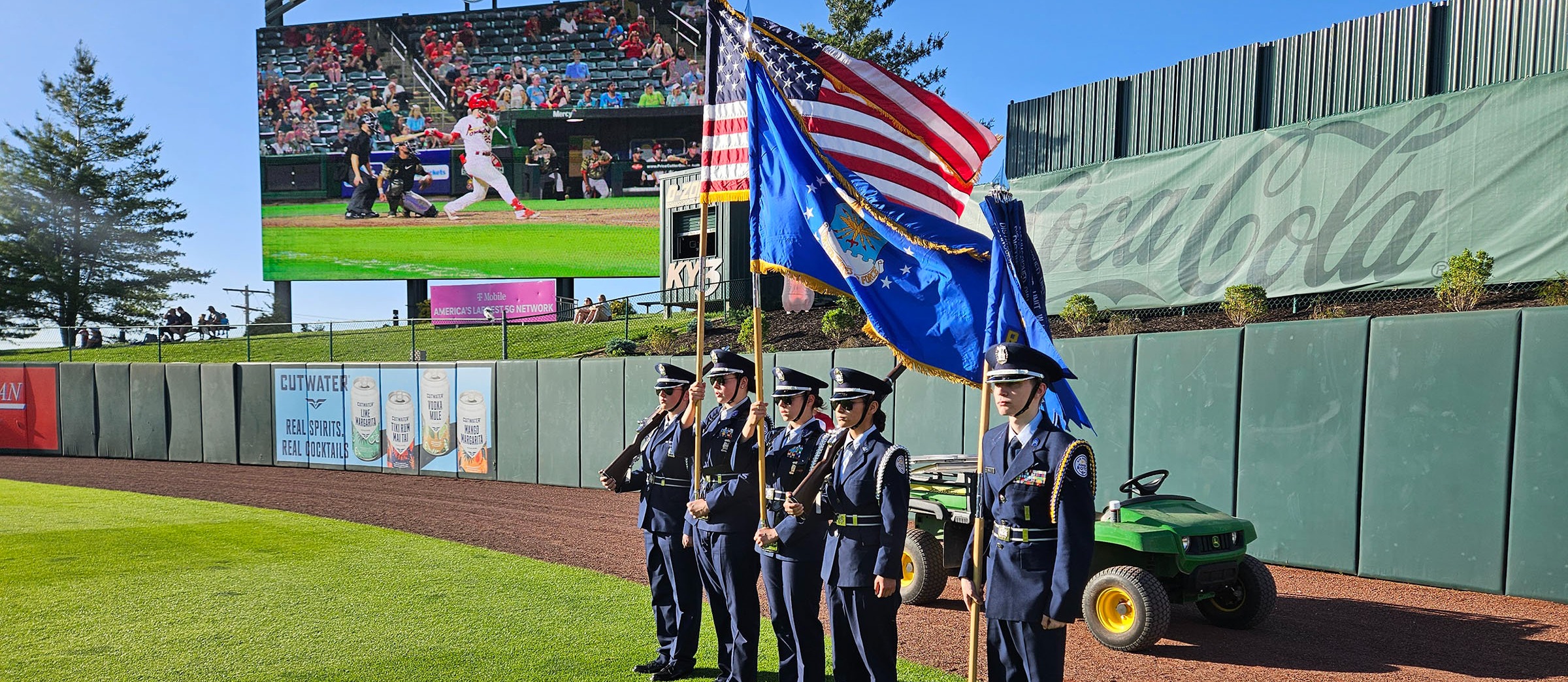 A five-member military color guard stands at attention on the outfield grass along the warning track at Hammons Field during a sunny day. The members wear dark blue dress uniforms, cap hats, and white gloves, with the center members holding the American flag and organizational flags. In the background, the stadium's large jumbotron displays a baseball game beneath the red "HAMMONS FIELD" sign.