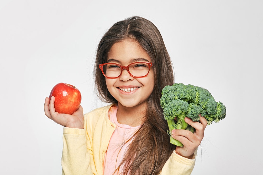 A girl in glasses holding an apple and a stalk of broccoli