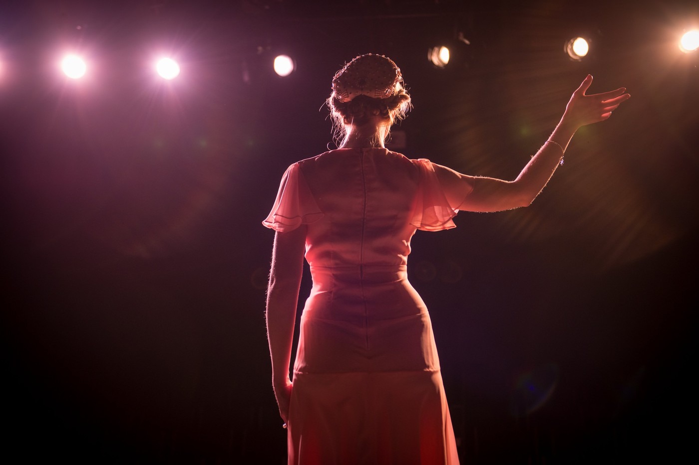 Woman's back, standing on stage with bright lights, her hand raised to the sky