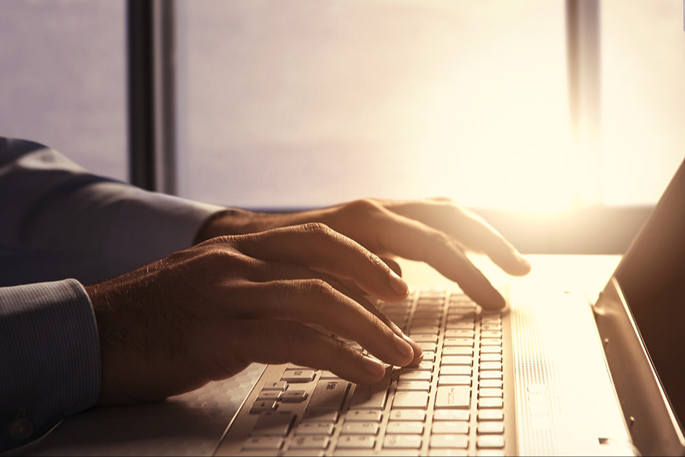 Hands typing on a laptop keyboard with sun glare in background