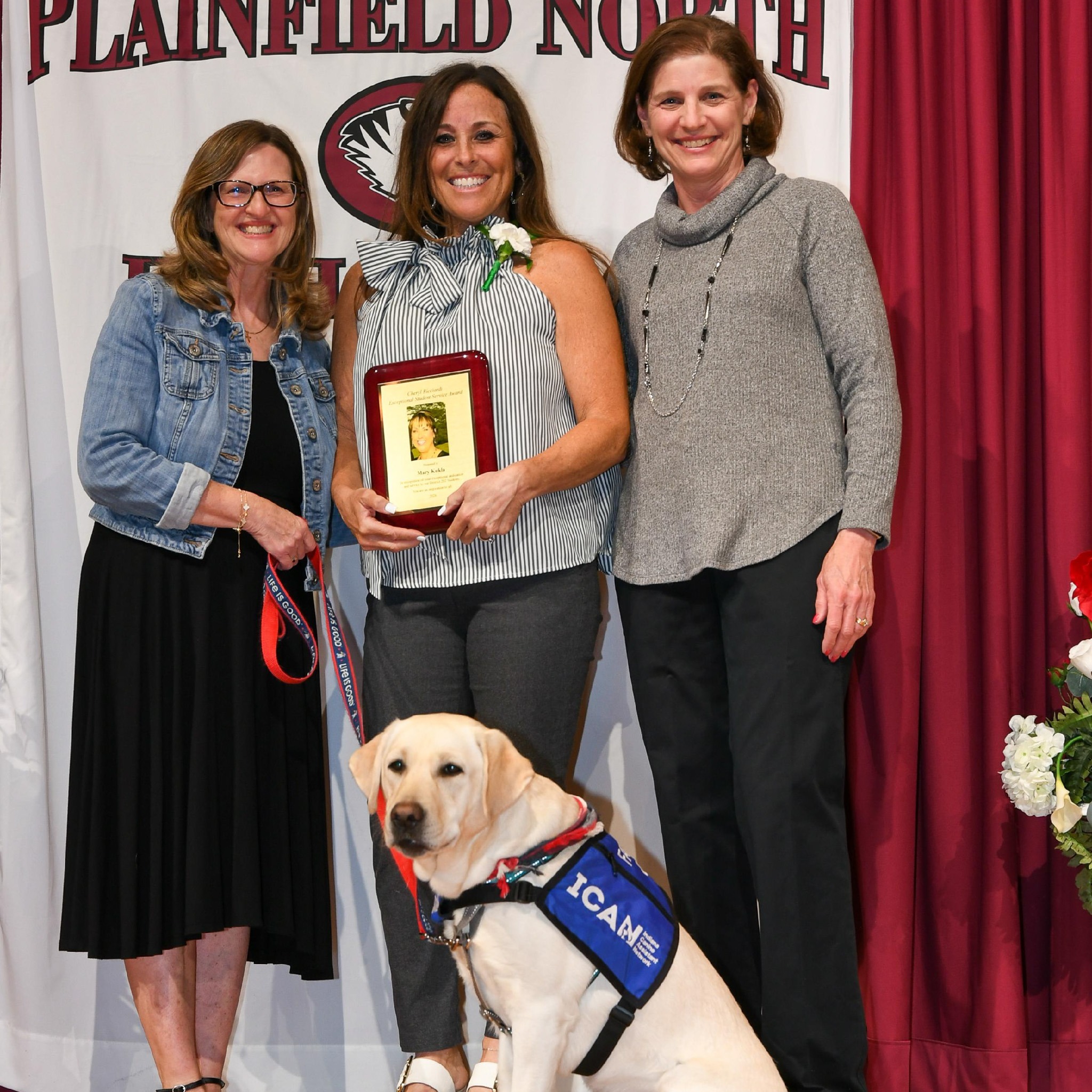 three woman and a dog stand in front of Plainfield North High School banner. Woman in middle hold awards plaque