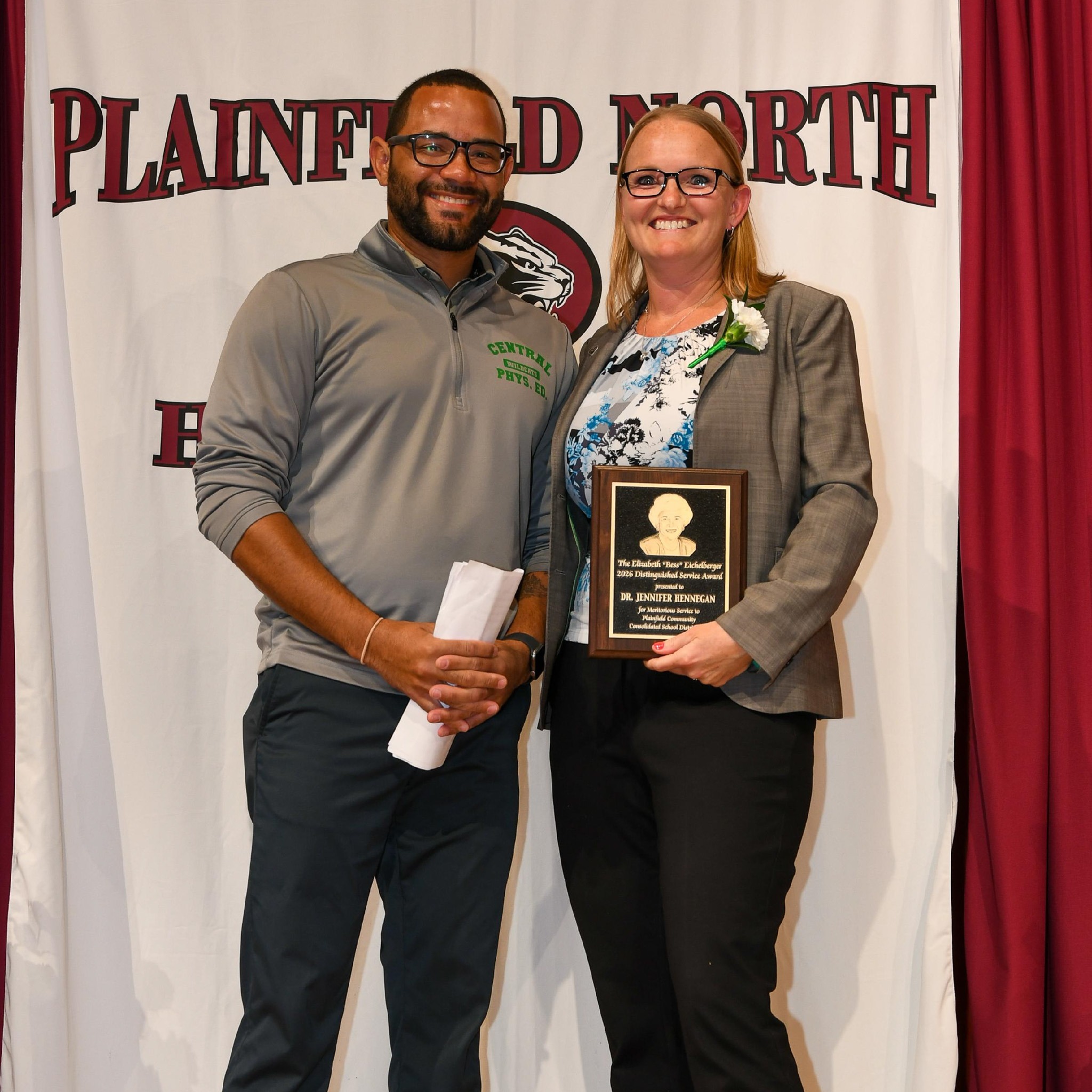 man and woman stand in front of Plainfield North banner. Woman on right holds awards plaque