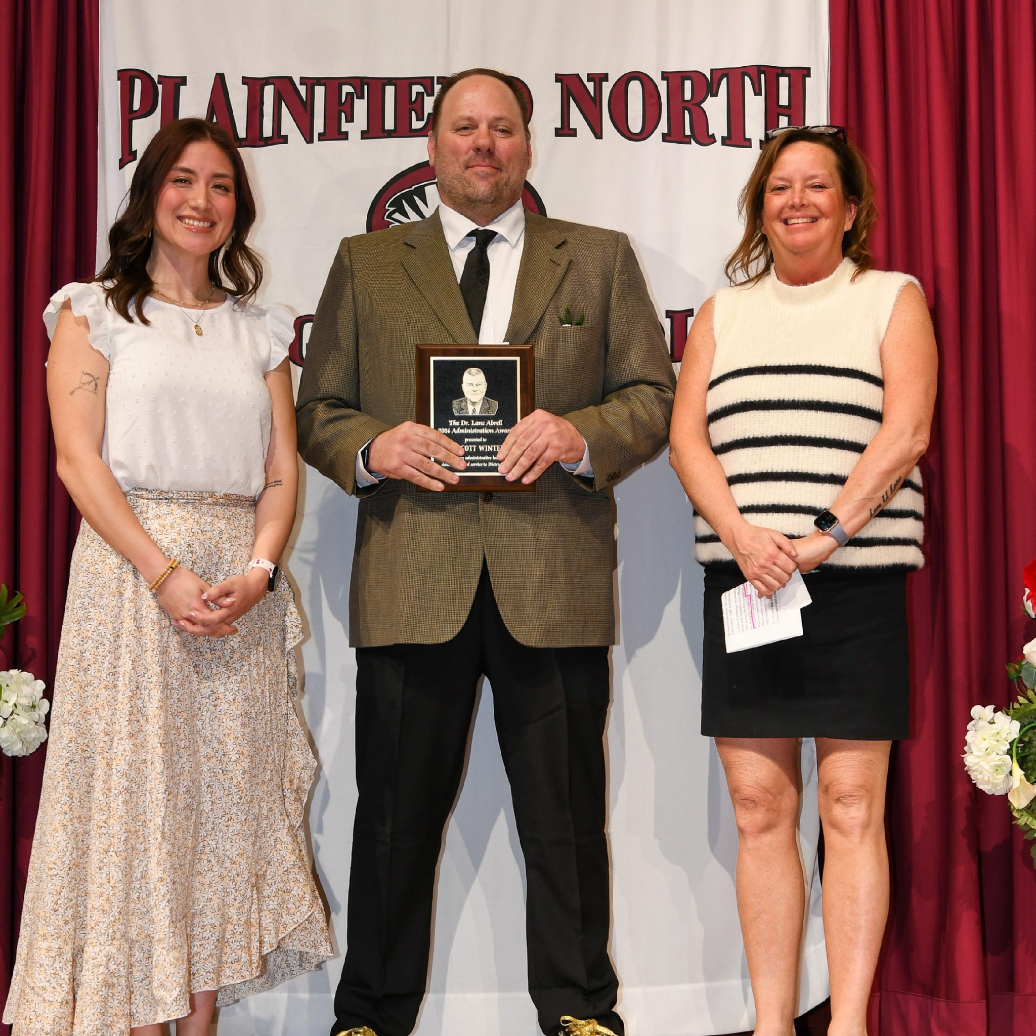 two woman and a man stand in front of Plainfield North banner. Man holds awards plaque.