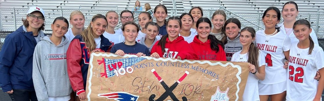 Lenape Valley field hockey player Kaidyn Gomez celebrates reaching 100 career points surrounded by teammates and coaches. The team holds a handmade sign recognizing Gomez and her 100-point milestone.