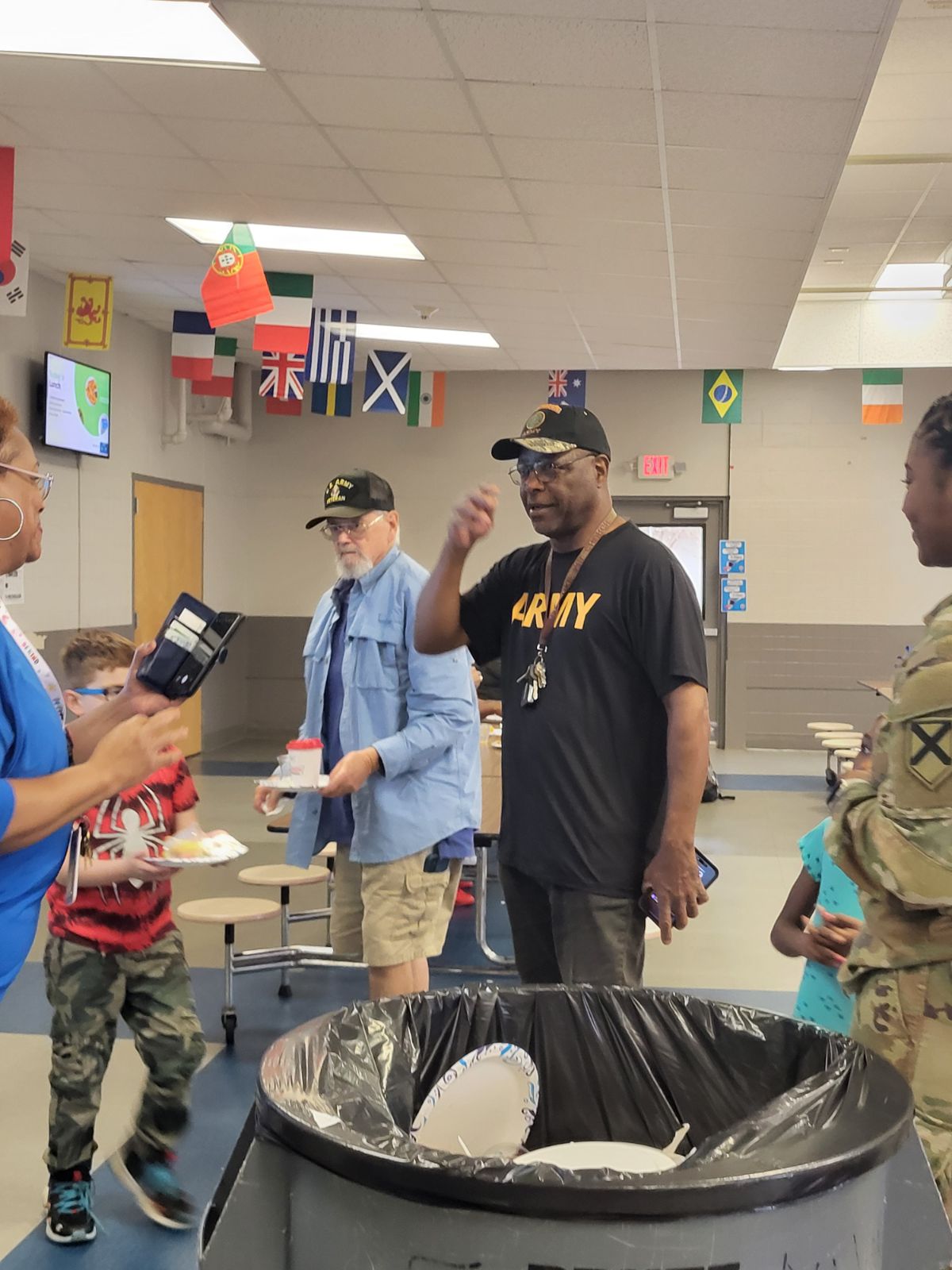Three military people wearing military gear with caps and black t-shirt , one teacher wearing blue, and two elementary children the cafeteria talking by a trashcan.